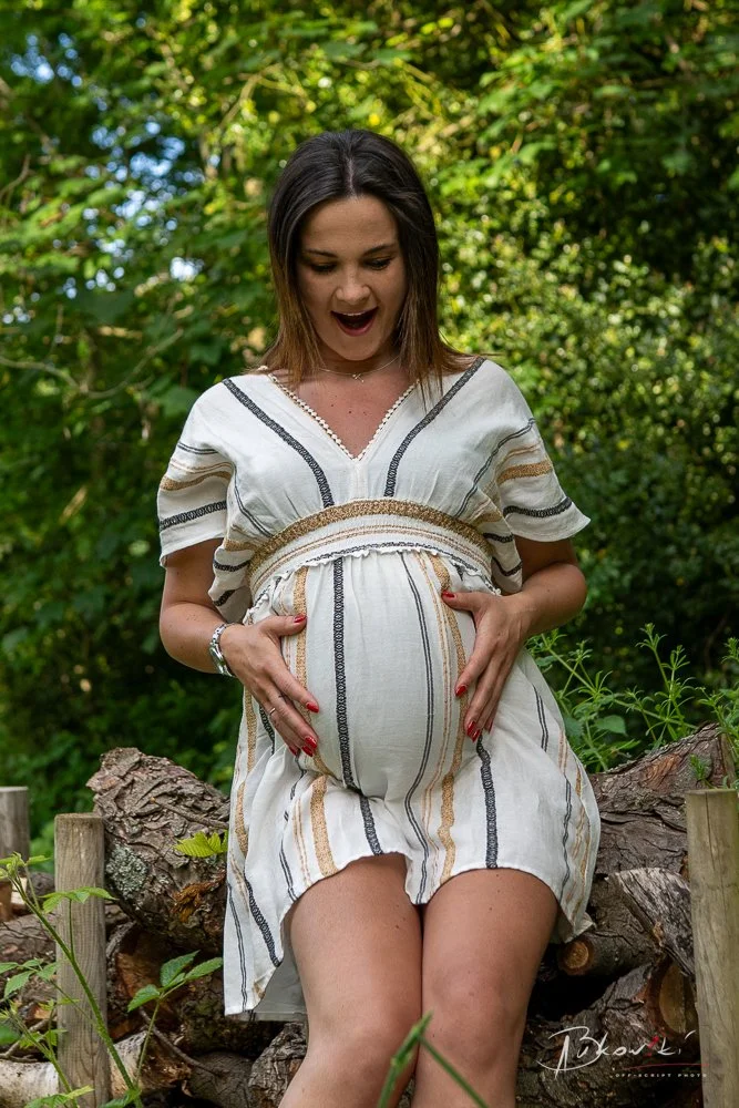 A pregnant woman sitting on a log outdoors, smiling and touching her belly, surrounded by green foliage.