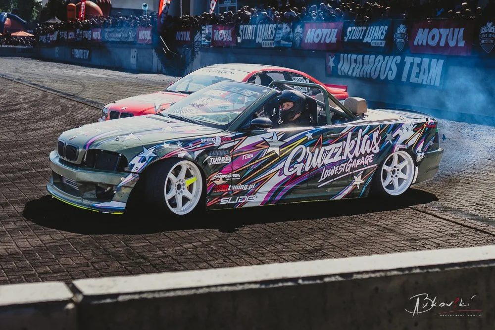 A drift car with a colorful, star-themed wrap and graffiti-style text 'Guzeklas Low St Garage' on the side, drifting on a racetrack with smoke and another red car in the background.