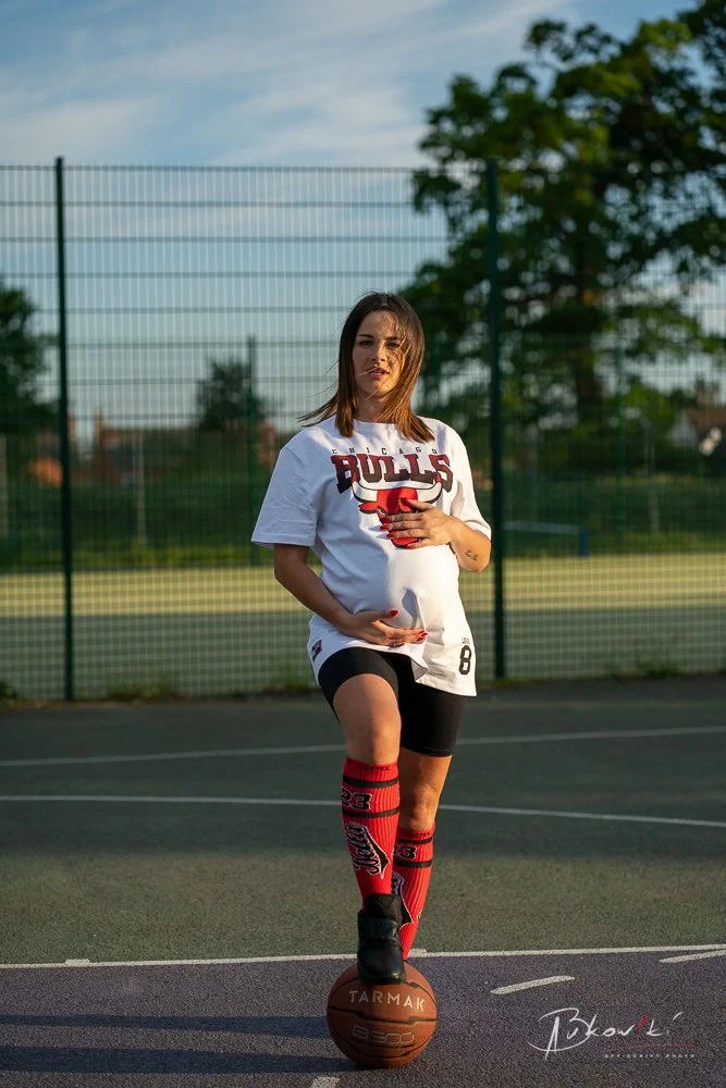 A woman in a Chicago Bulls T-shirt and black shorts balancing on one leg with her foot on a basketball on an outdoor court, with a fence and trees in the background.
