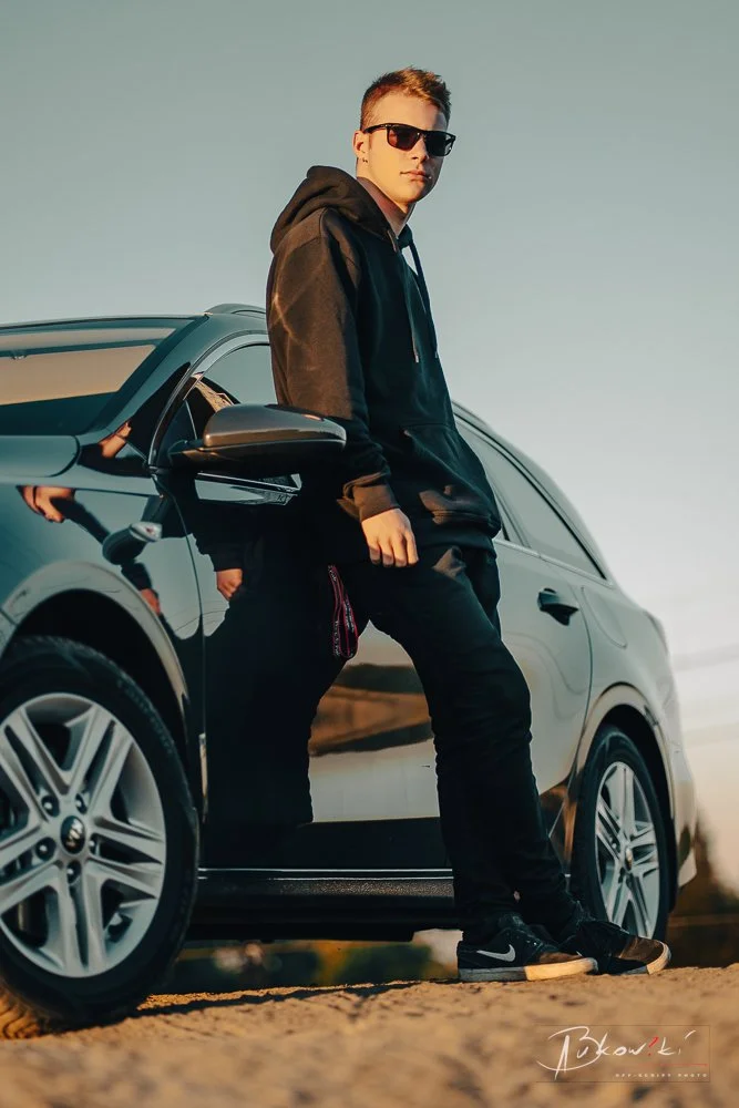 A young man wearing sunglasses, a black hoodie, and black pants leaning against a black car outdoors during sunset.