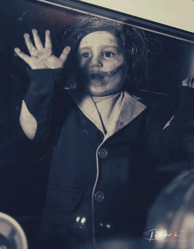A young child with a bruised face, lying on a glass surface, looking upwards with wide eyes and hand pressed against the glass, wearing a dark jacket over a shirt.