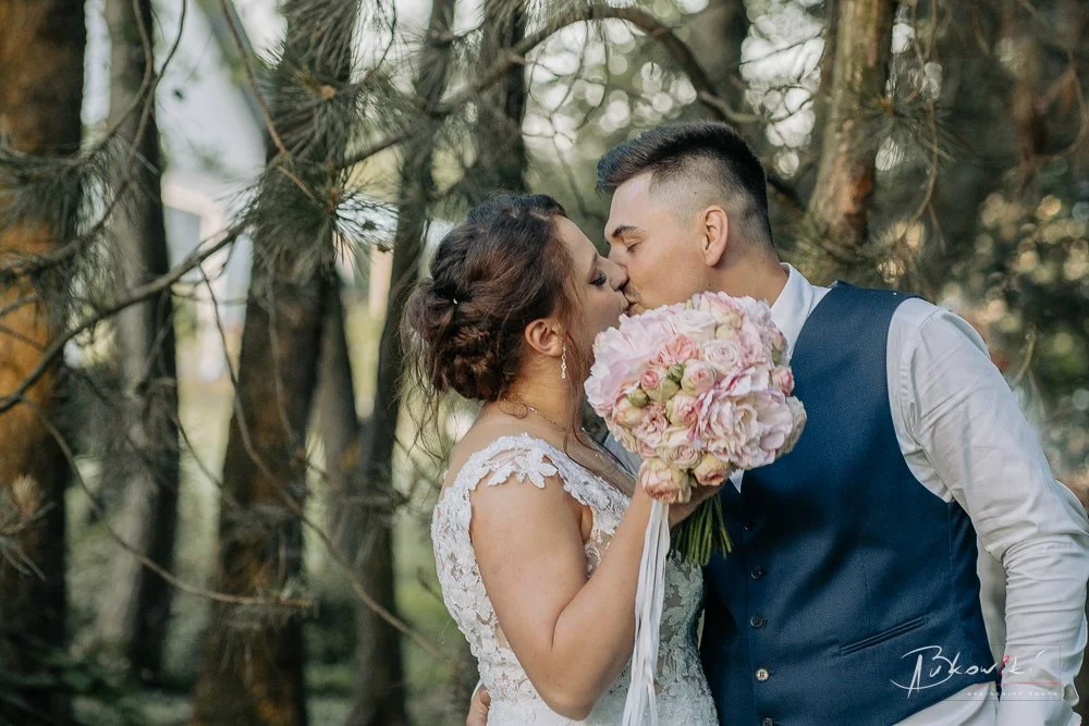A bride and groom kissing outdoors, with the bride holding a pink bouquet of flowers, surrounded by trees.