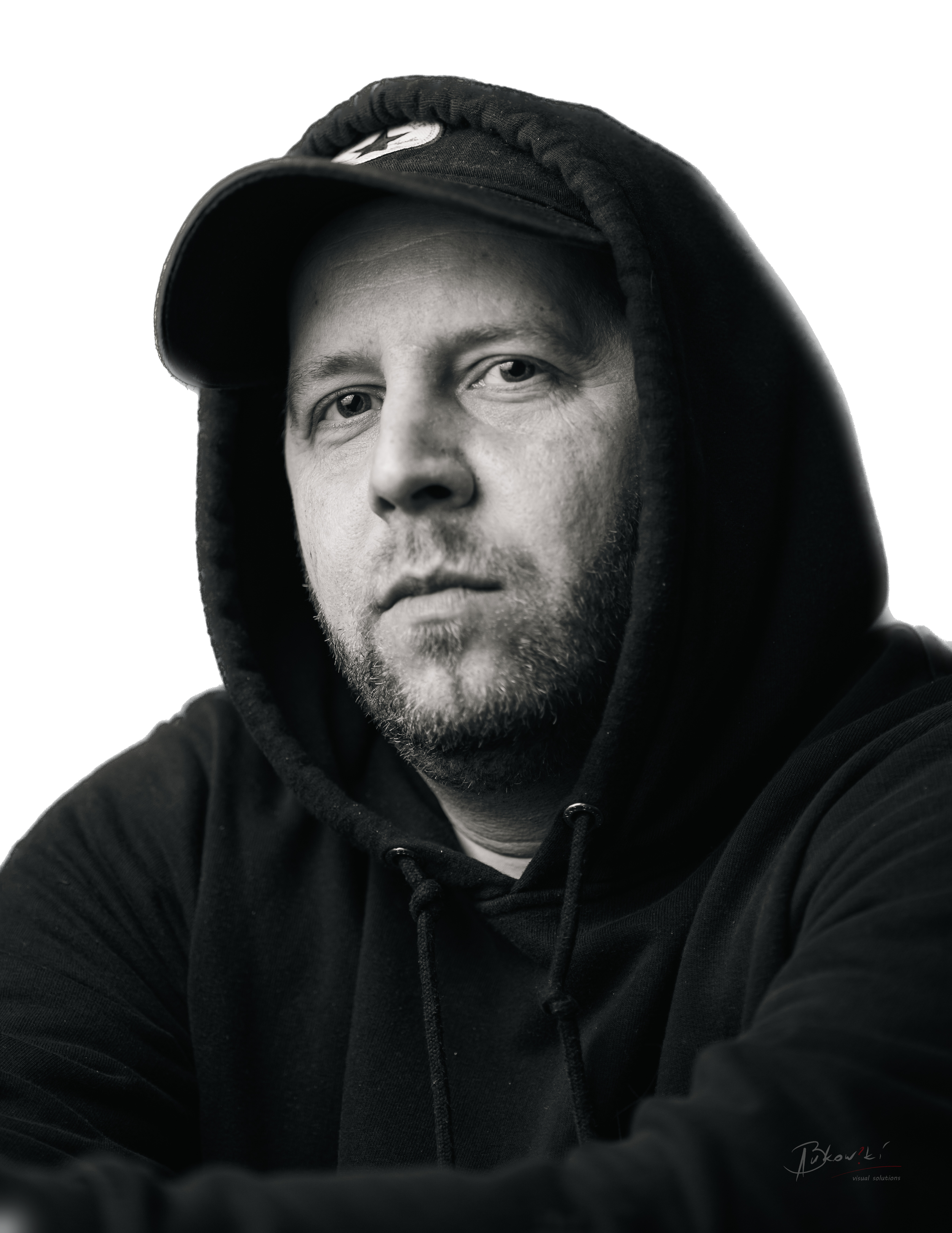 Close-up black and white portrait of a man wearing a black hoodie and a black cap, looking directly at the camera with a serious expression.