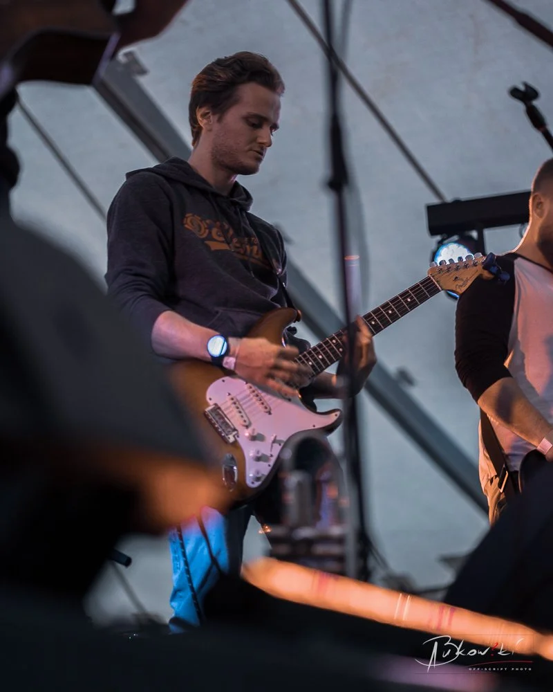 Young man with brown hair playing electric guitar on stage during a live music performance, with stage equipment and other band members visible.