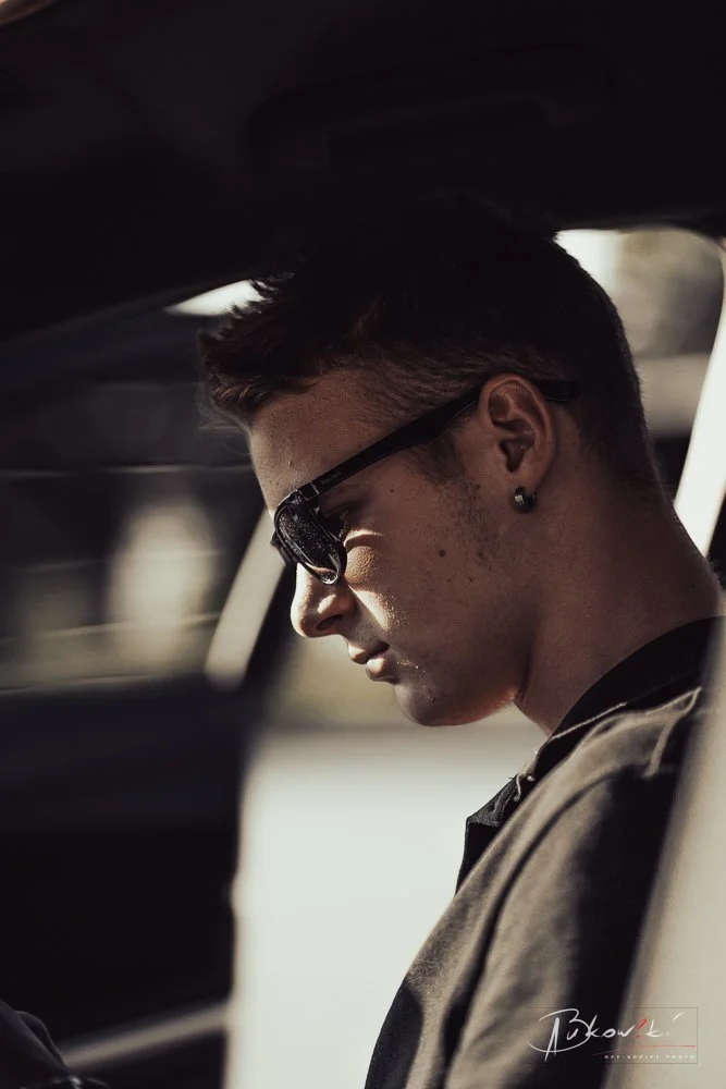 Side profile of a young man wearing sunglasses with short hair, earrings, and a dark shirt, sitting inside a vehicle.