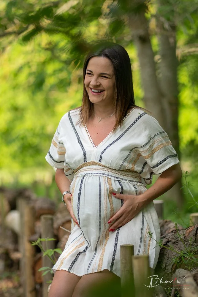 A pregnant woman with light skin and shoulder-length brown hair stands outdoors in a lush green forest, smiling and cradling her baby bump with her hands, wearing a white patterned dress.