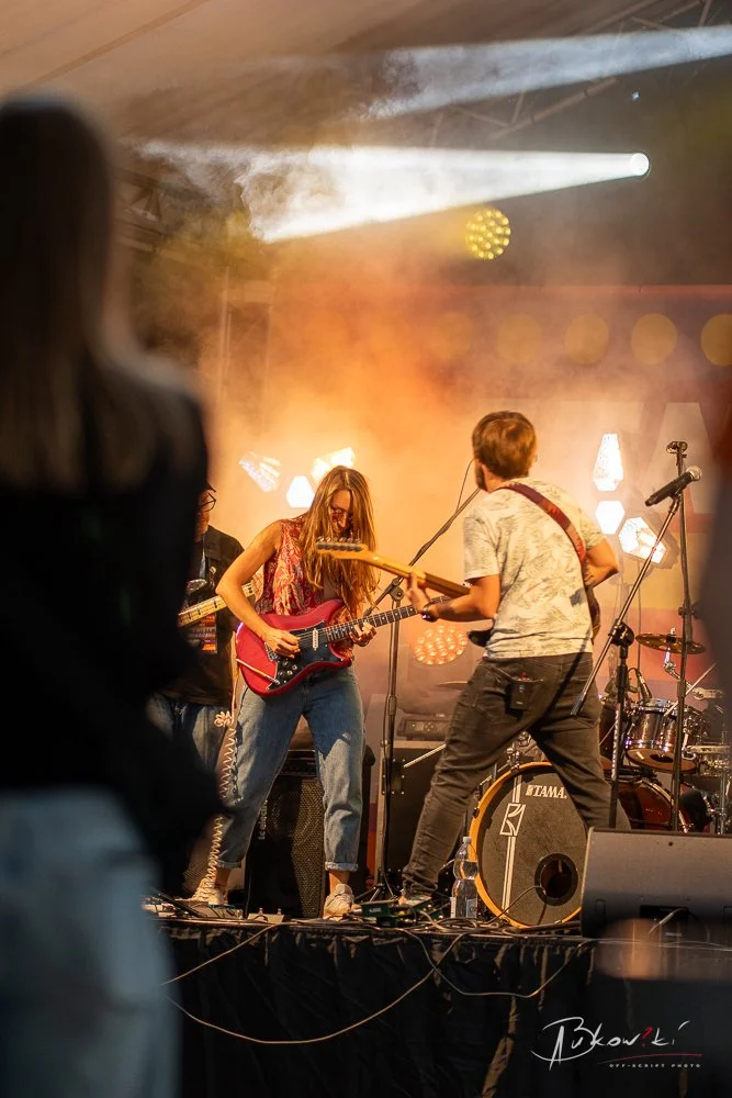 A live band performance with a woman playing an electric guitar on stage, surrounded by stage lights and fog.