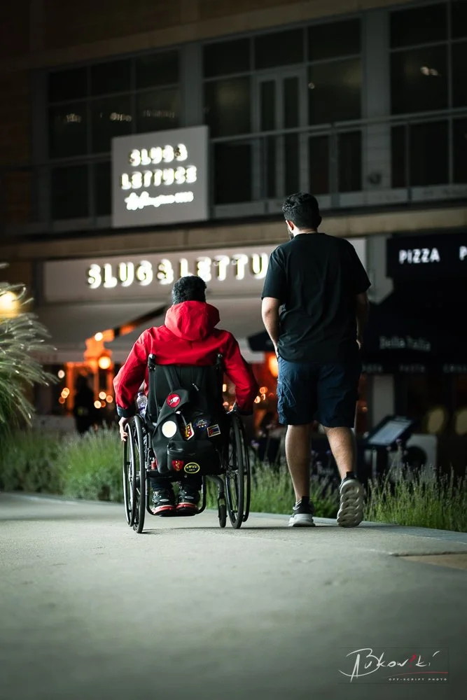 Two men walking on a sidewalk at night, one in a wheelchair and the other beside him, outside a restaurant called Slugs Lettuce with bright illuminated signs.