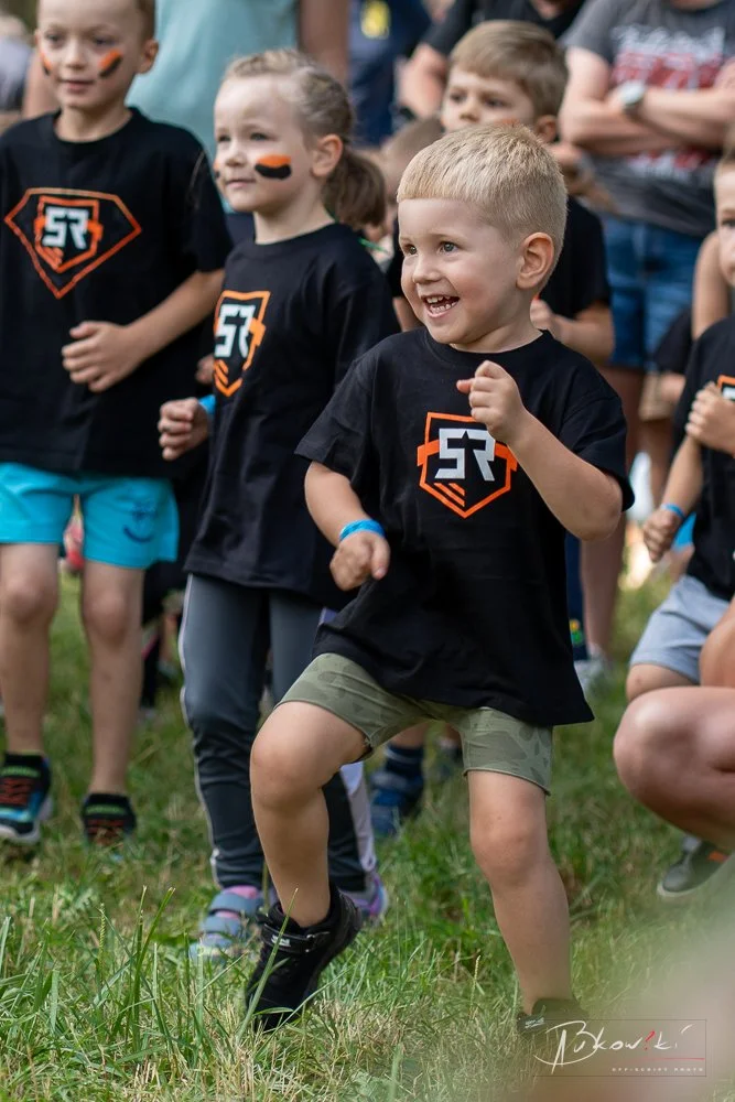 Children participating in an outdoor event, wearing matching black t-shirts with a logo, one boy in the front smiling and dancing on grass.