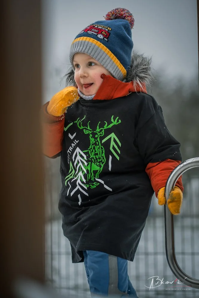 A young boy dressed in winter clothing, including a colorful beanie, orange gloves, and a black jacket with a green deer graphic, stands outdoors near a metal fence on a cold day with a blurry background.