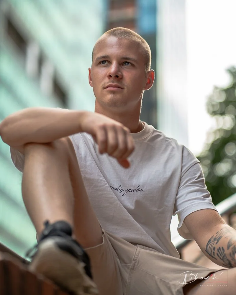 Young man with a shaved head sitting outdoors, looking thoughtfully into the distance, wearing a white t-shirt and beige shorts.