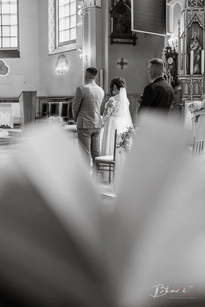 A wedding ceremony with a bride and groom standing at the altar inside a church, with two men nearby. The bride is wearing a white gown with lace, and the groom in a light suit. The church features stained glass windows and religious statues, with th