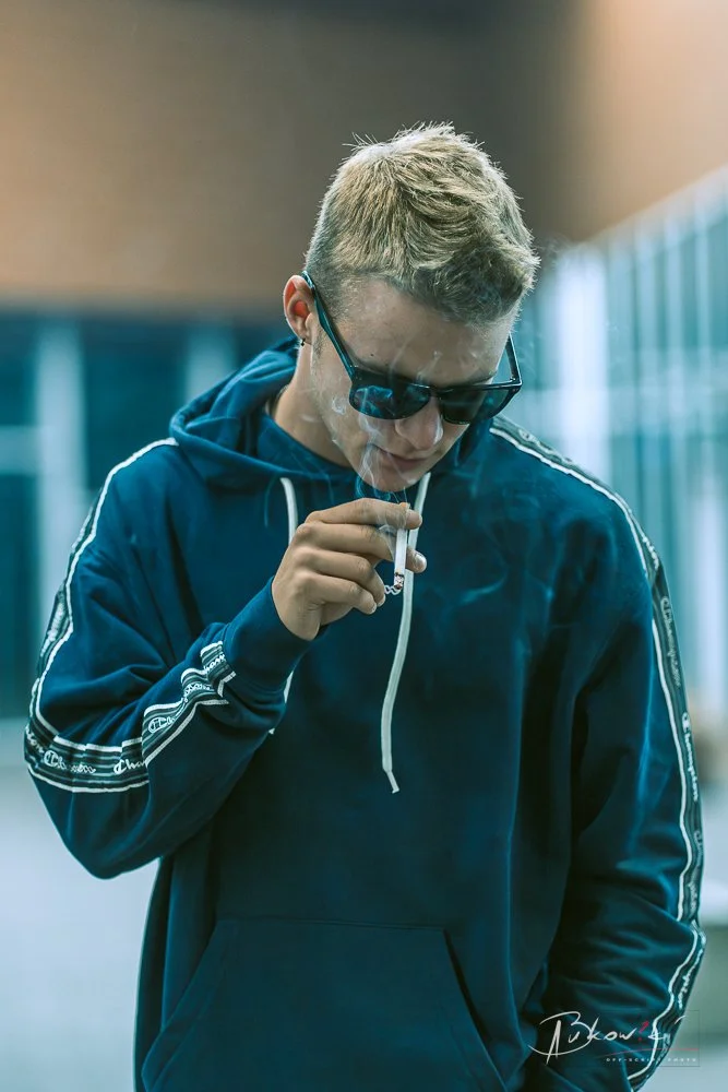 Young man with blond hair and sunglasses smoking a cigarette outdoors.
