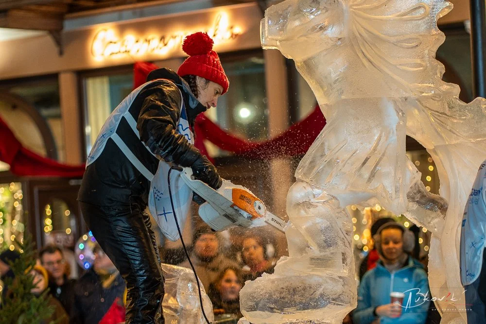 Person wearing a red knit hat and black jacket carving ice sculpture at a holiday event with onlookers in the background.