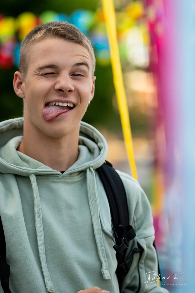 Young man sticking out his tongue and smiling while winking at the camera, wearing a light green hoodie and black backpack, with colorful blurred background.
