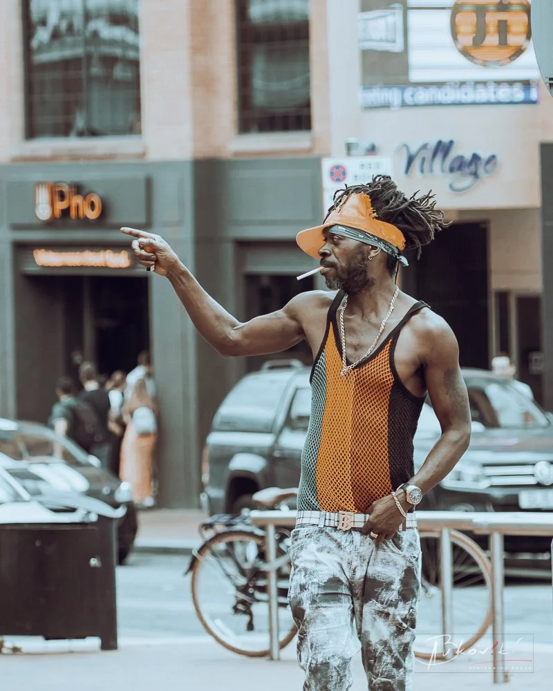 A man with dreadlocks wearing an orange visor, sunglasses, a black and orange mesh tank top, and patterned pants, standing on a city street pointing.