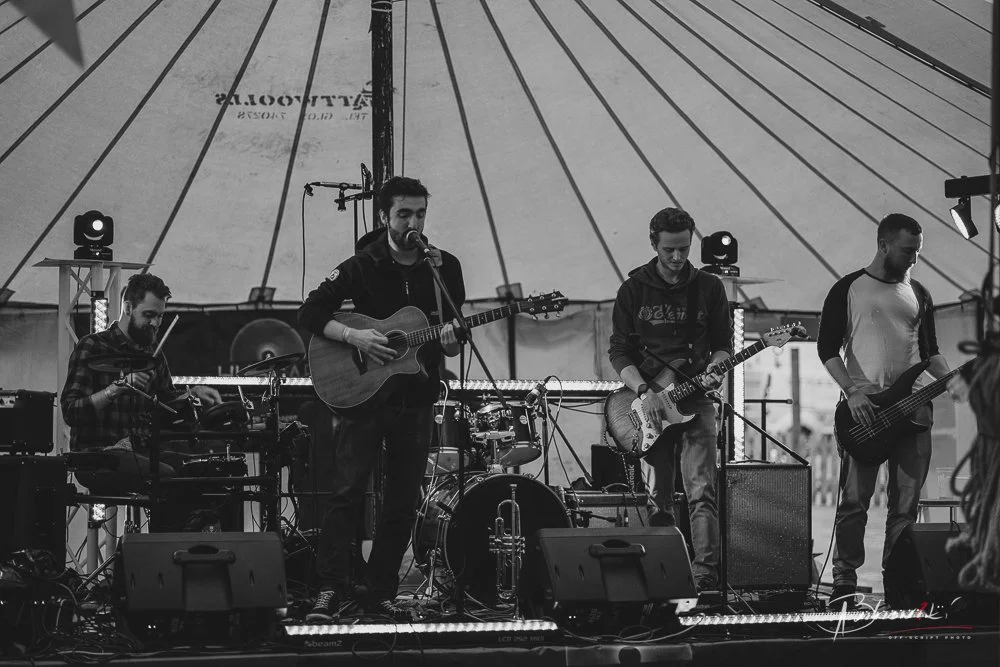 A band performing on stage inside a tent, with four members playing musical instruments and singing.