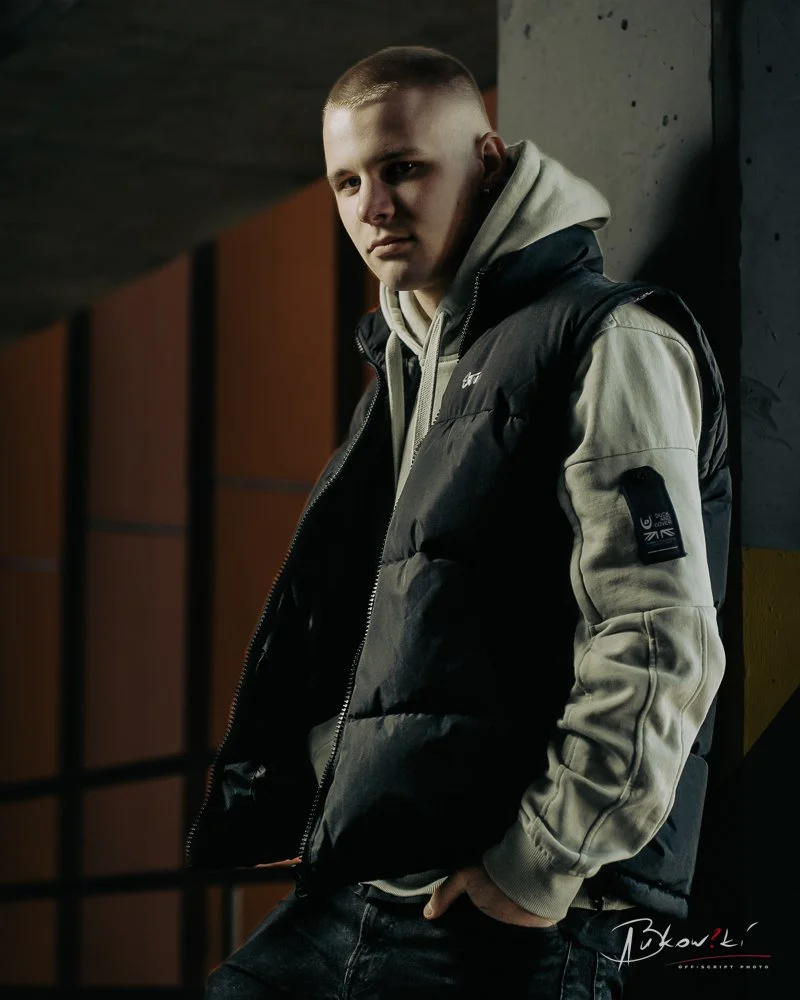 Young man with a buzz cut hairstyle leaning against a concrete wall in an indoor setting, wearing a black and beige puffer jacket and a hoodie, with serious facial expression.