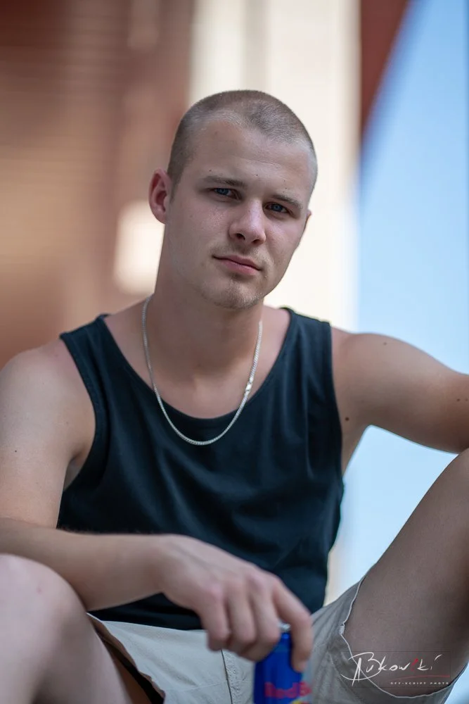 A young man with a shaved head wearing a black sleeveless shirt and a silver chain necklace, sitting outdoors with a serious expression.