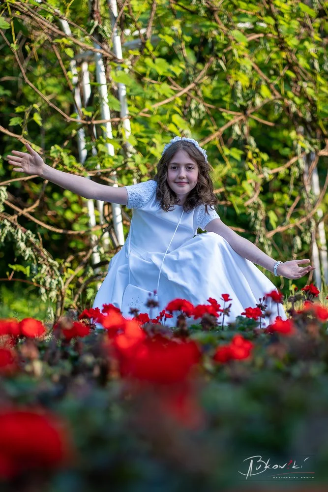 A young girl in a white dress and headband is outdoors, sitting amid red flowers with her arms outstretched, in front of green foliage.