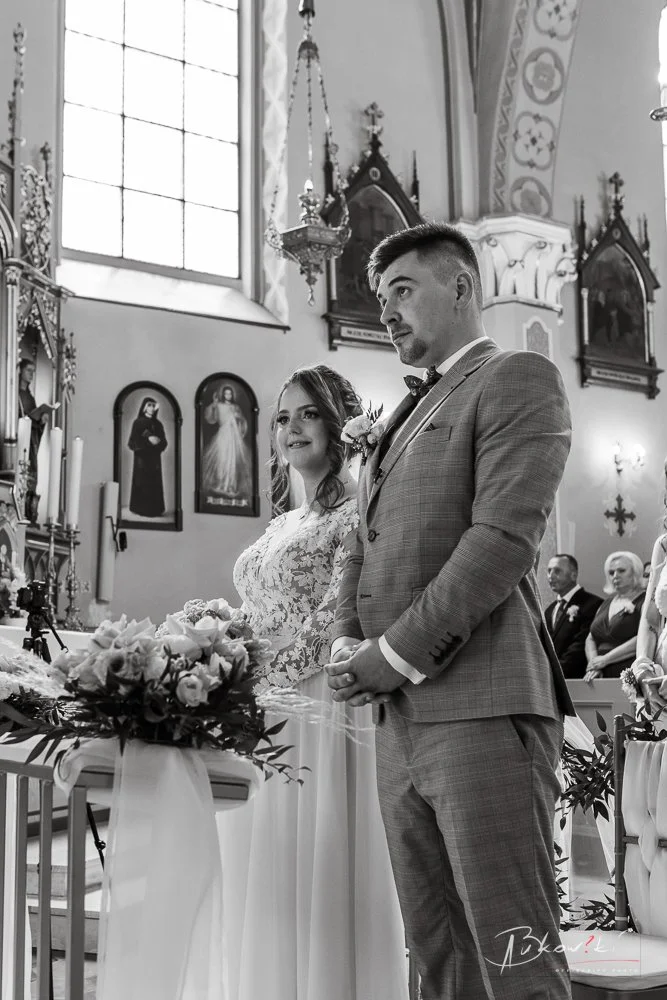 A bride and groom standing during a wedding ceremony inside a church, with wedding guests in the background and religious paintings on the walls.
