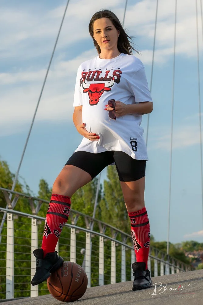 Pregnant woman wearing Chicago Bulls jersey, shorts, and soccer socks, standing on a bridge with one foot on a basketball, holding sunglasses, outdoors with blue sky and trees in the background.