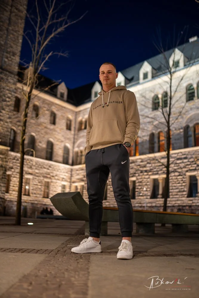 Young man in beige Tommy Hilfiger hoodie, black Nike joggers, and white sneakers standing outdoors at night with historic stone building and leafless trees in background.