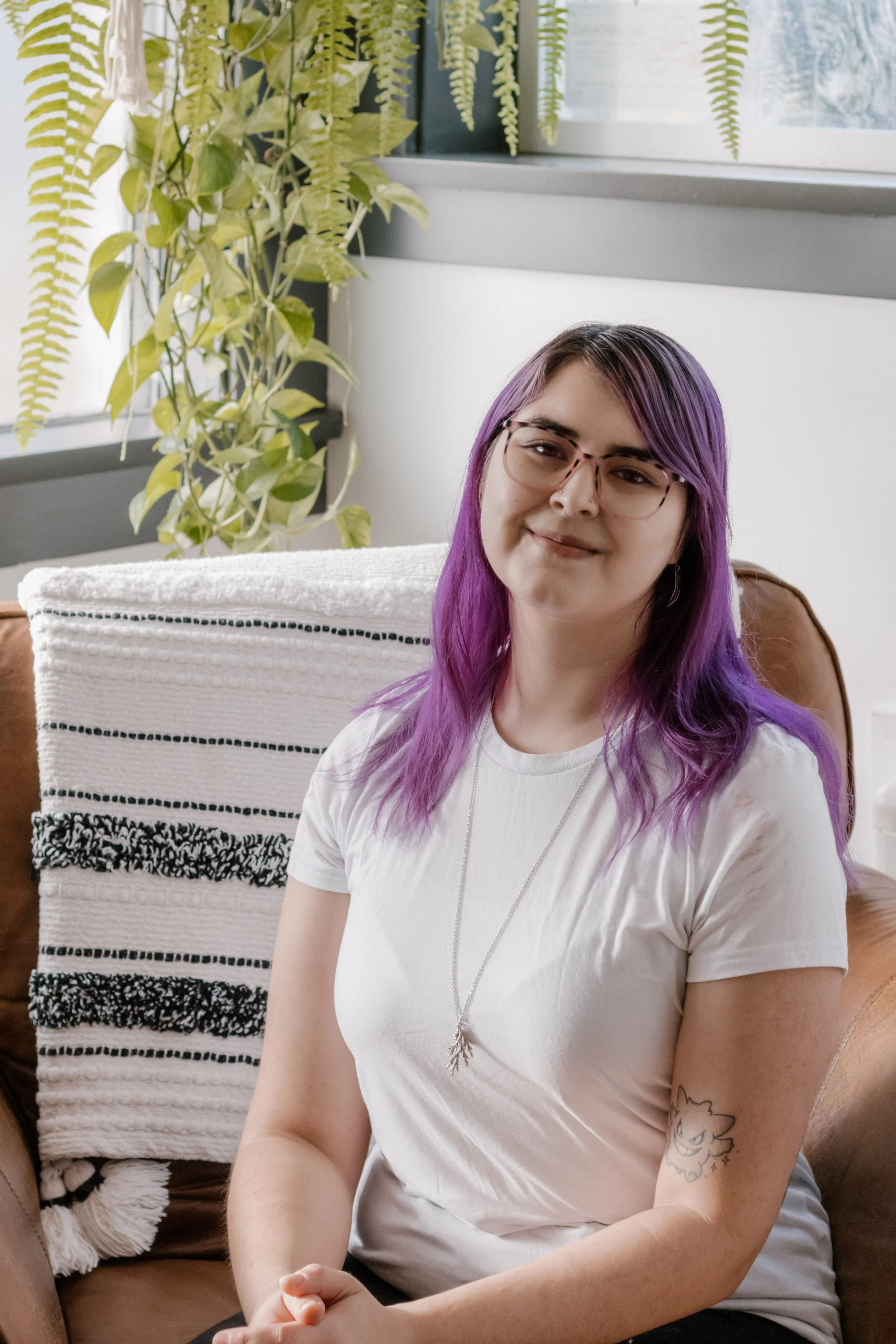 A young woman with purple hair, glasses, and a tattoo on her arm, sitting in a brown leather chair with a white and black patterned blanket behind her, in a well-lit room with potted plants and windows.