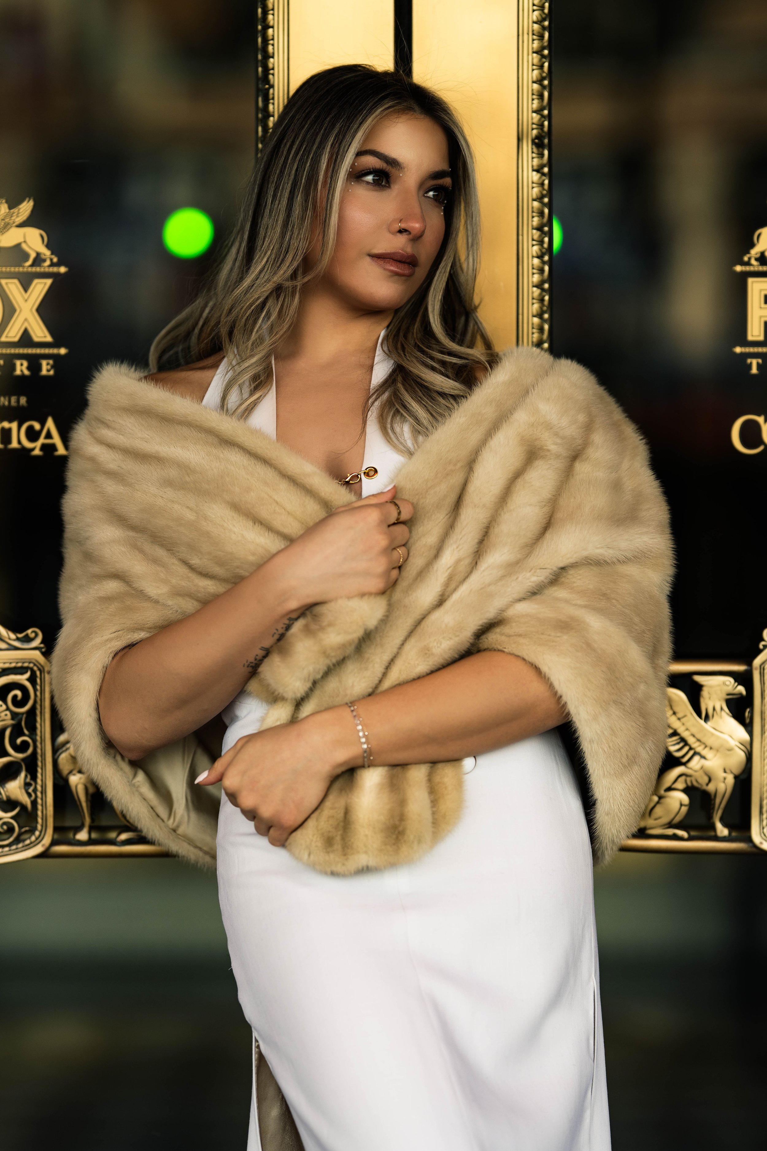 A woman with blonde hair stands in front of a black and gold background, wearing a white dress and a beige fur stole, looking to the side.