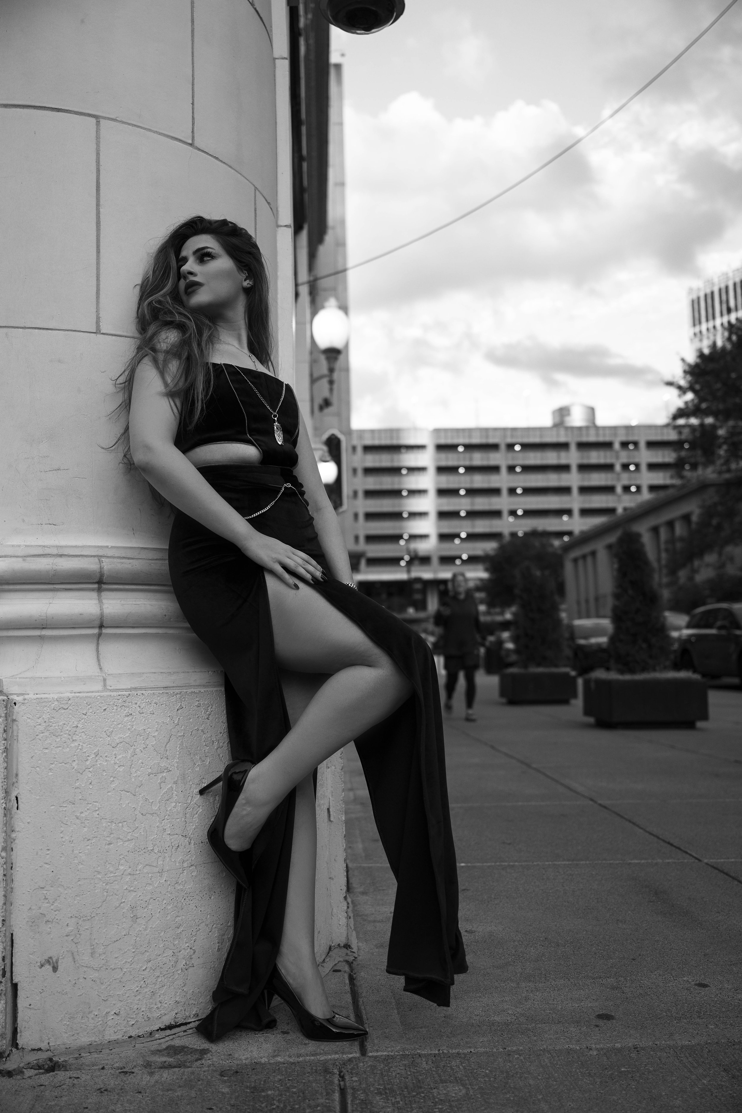 A woman in a long black dress sitting against a large column on a city sidewalk, with buildings and a cloudy sky in the background.
