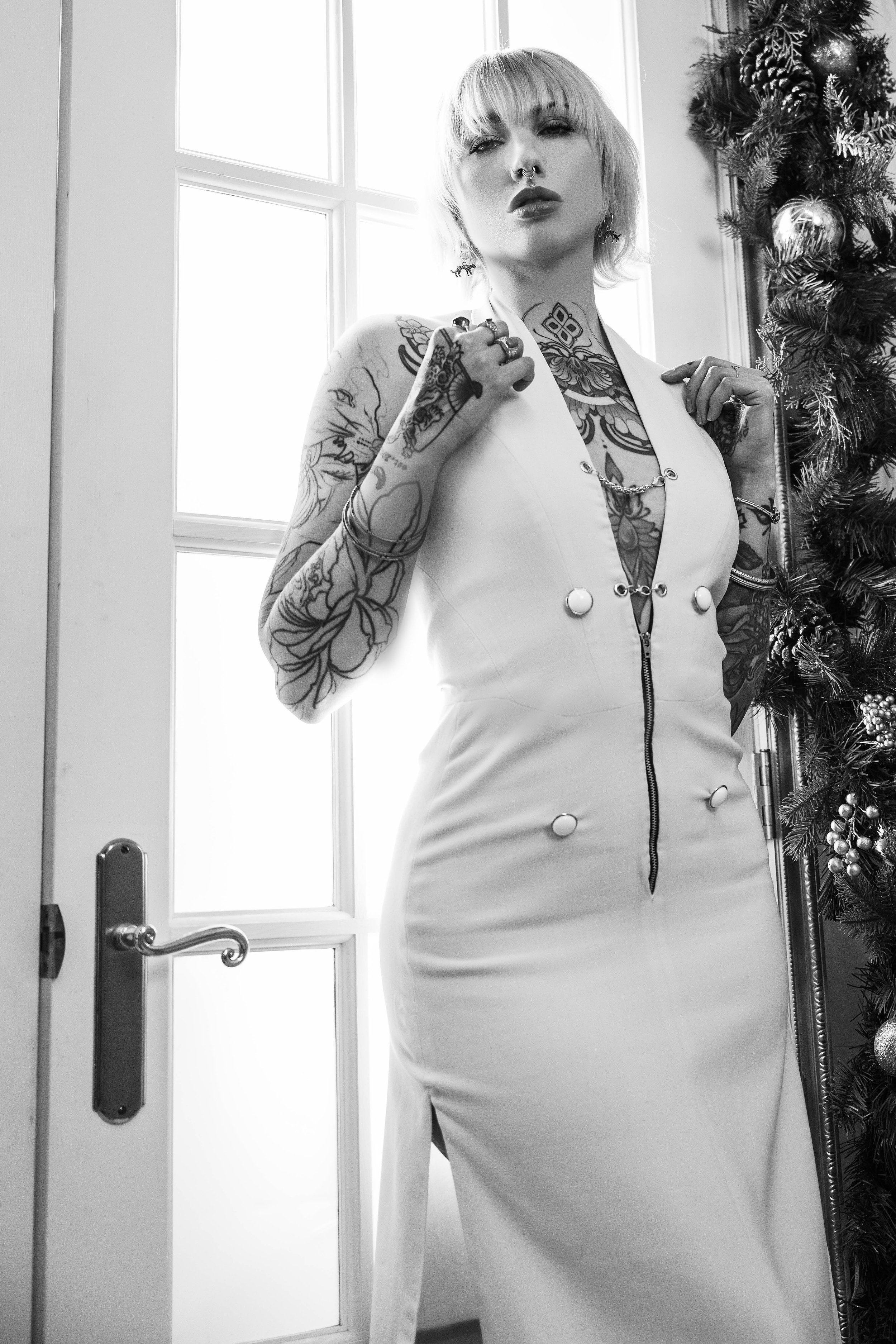 Black and white photo of a woman with tattoos, short hair, and a septum ring, standing in front of a window and a Christmas tree, wearing a fitted sleeveless dress with a zipper front.