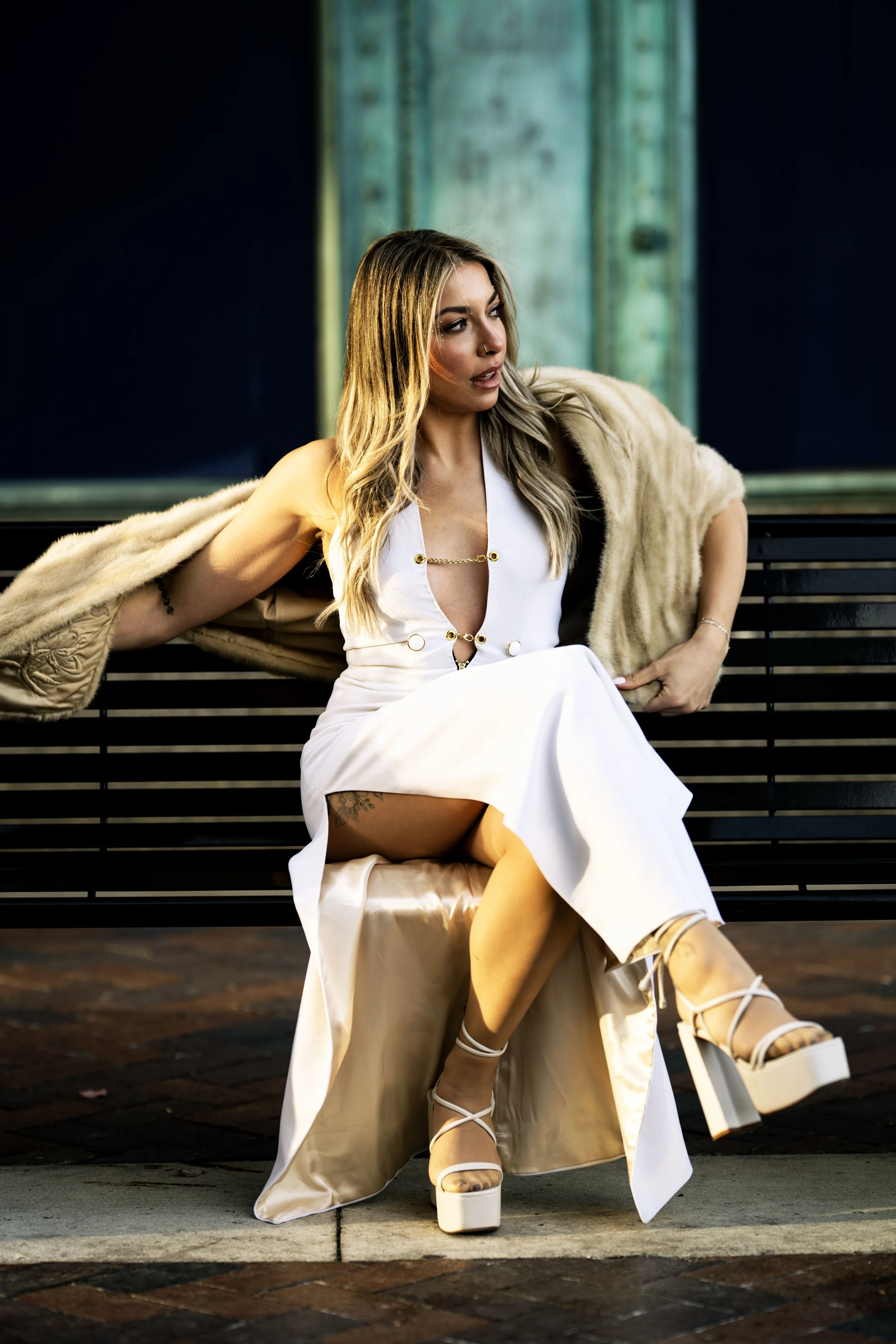 A woman sitting on a bench, wearing a white dress, platform heels, and a fur coat draped over her shoulders.