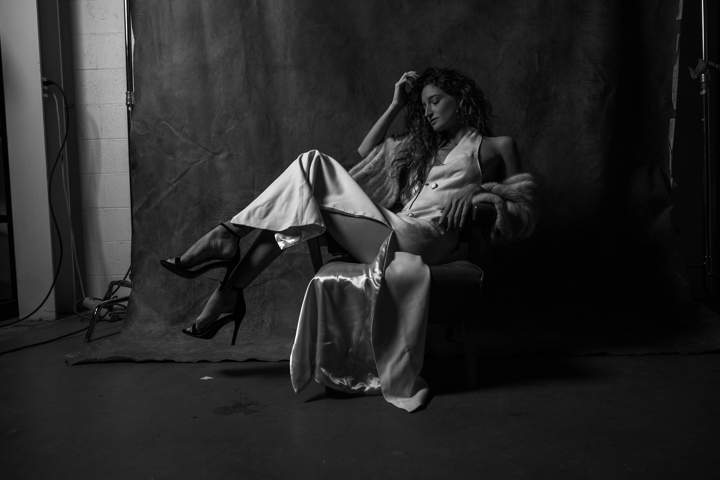 A woman with curly hair sitting on a chair, wearing a light-colored satin dress and high-heeled shoes, with a fur stole draped over her arm, in a black-and-white photo with a dark backdrop.
