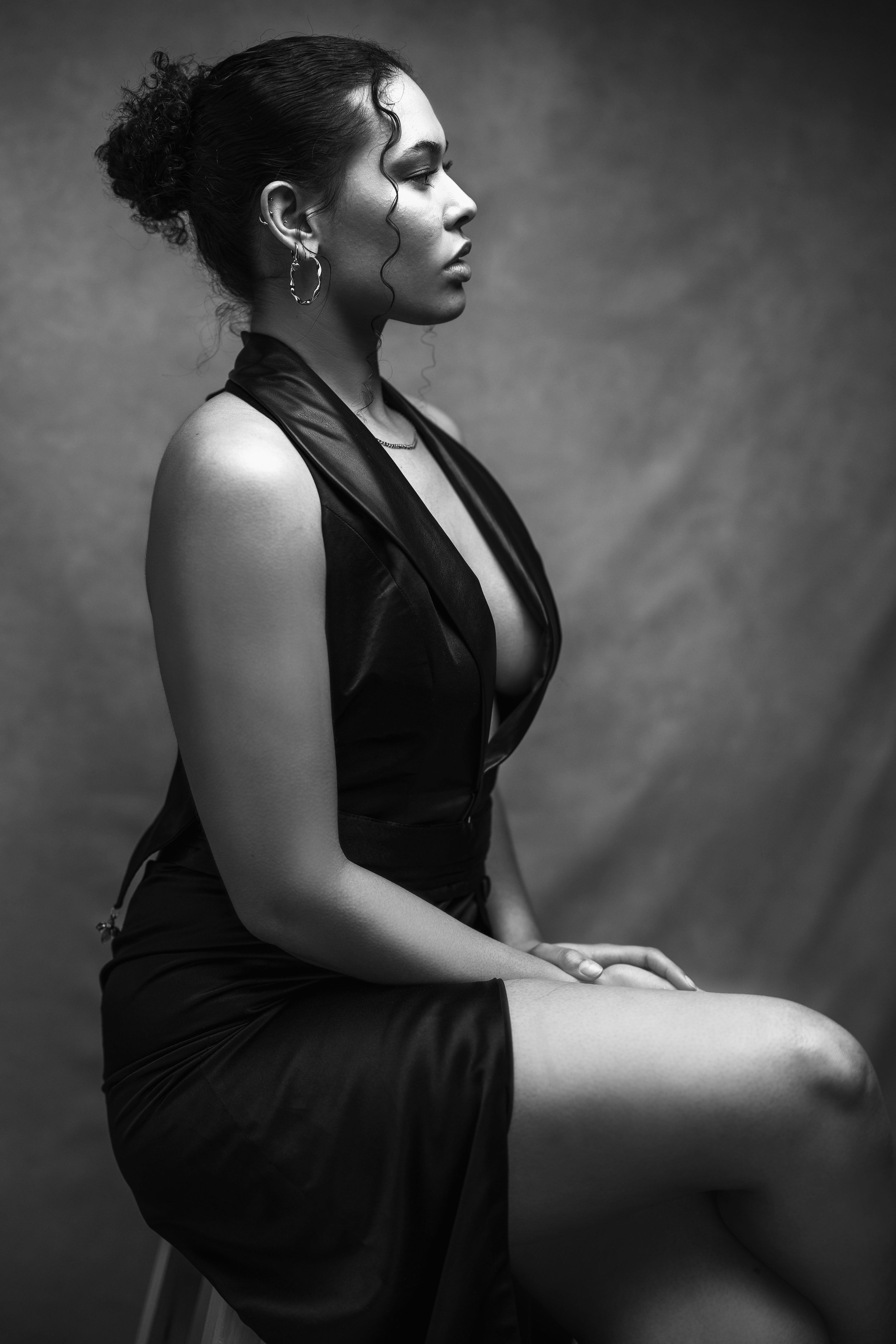 A black-and-white portrait of a woman in profile, sitting with her back straight and her hands resting on her lap. She has curly hair in a bun, wearing hoop earrings, a sleeveless leather vest, and a dress with a slit.