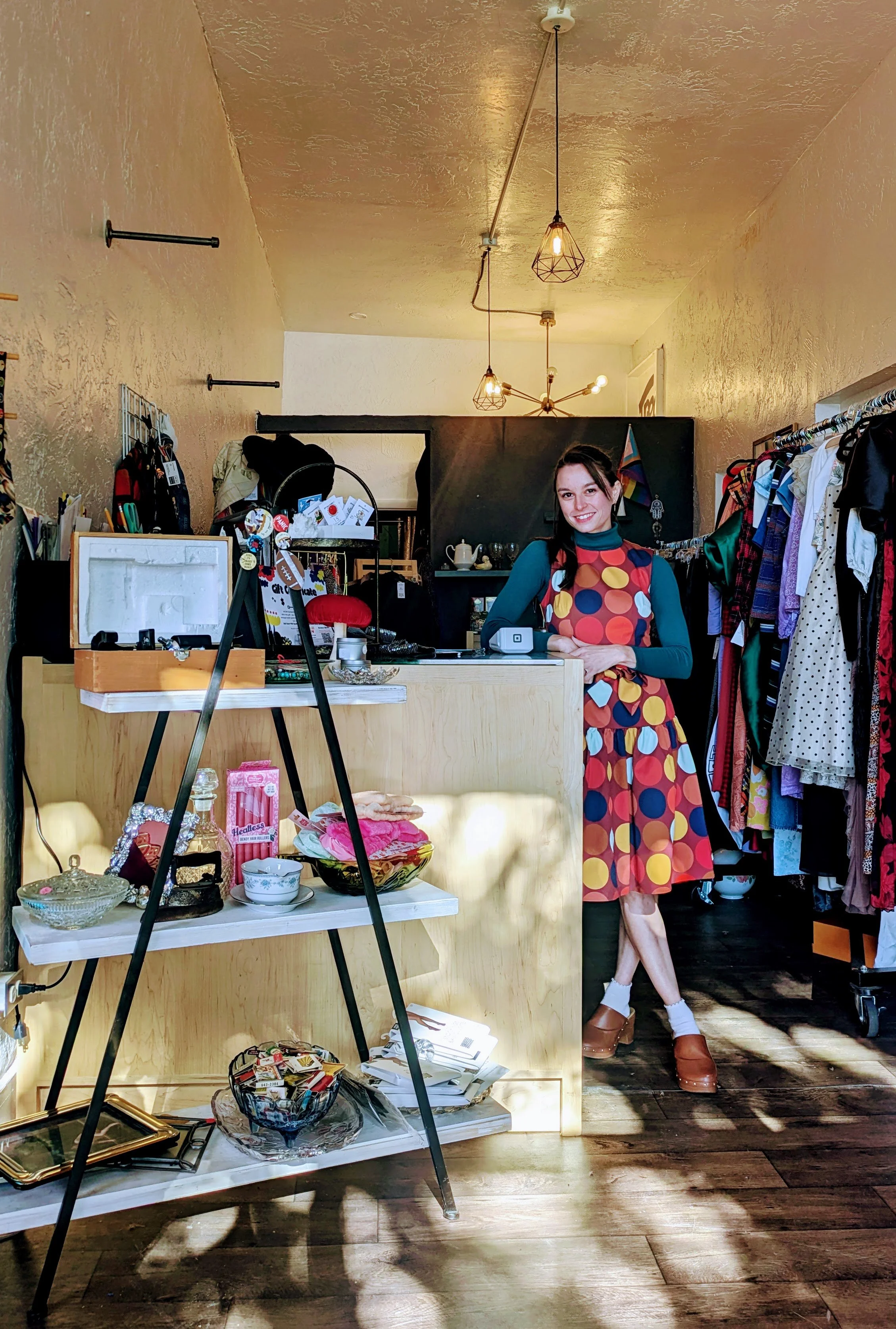 A woman standing behind a boutique store counter, wearing a colorful polka dot dress and brown platform shoes. The store has clothing on racks to her right and decorative items on a white shelf in front of her.