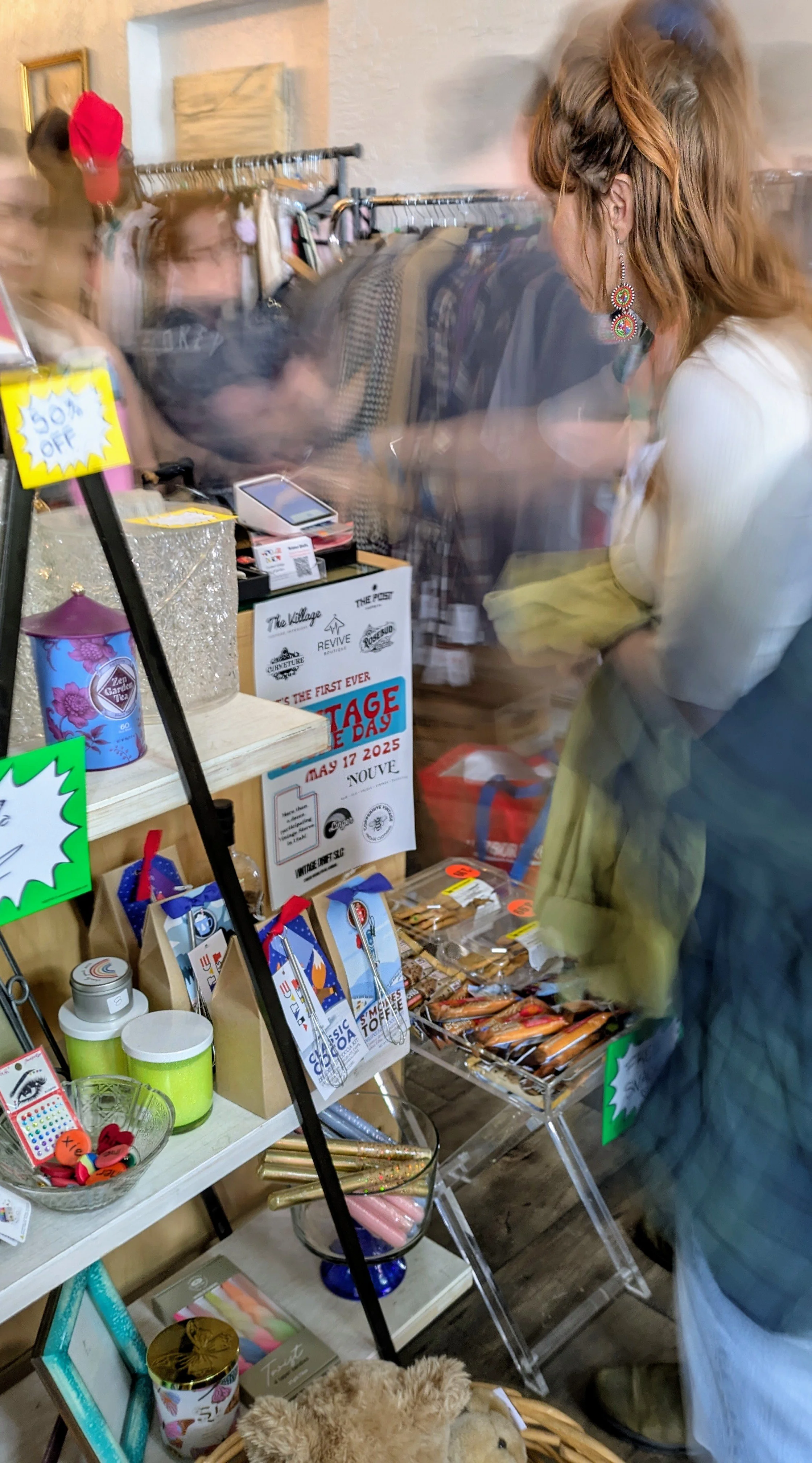 Indoor scene at a thrift or gift shop with shelves displaying candles, toys, and decorative items. A woman with this side profile is browsing. There is a sale sign and various merchandise on display.
