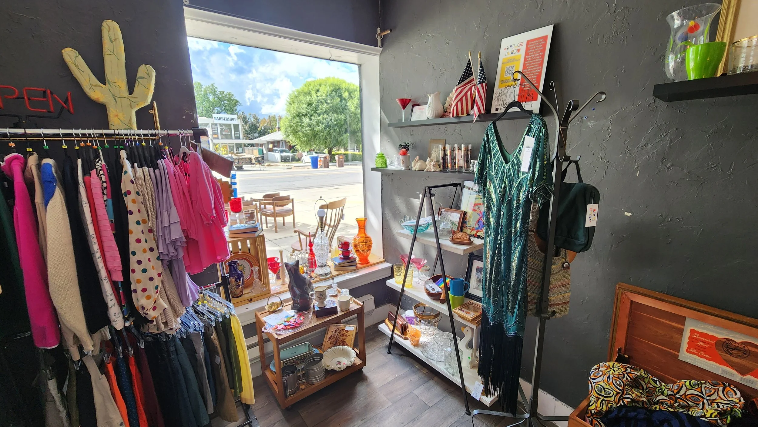 Interior of a thrift shop with clothing rack, glassware, and decorative items on shelves, facing a storefront window showing street outside.