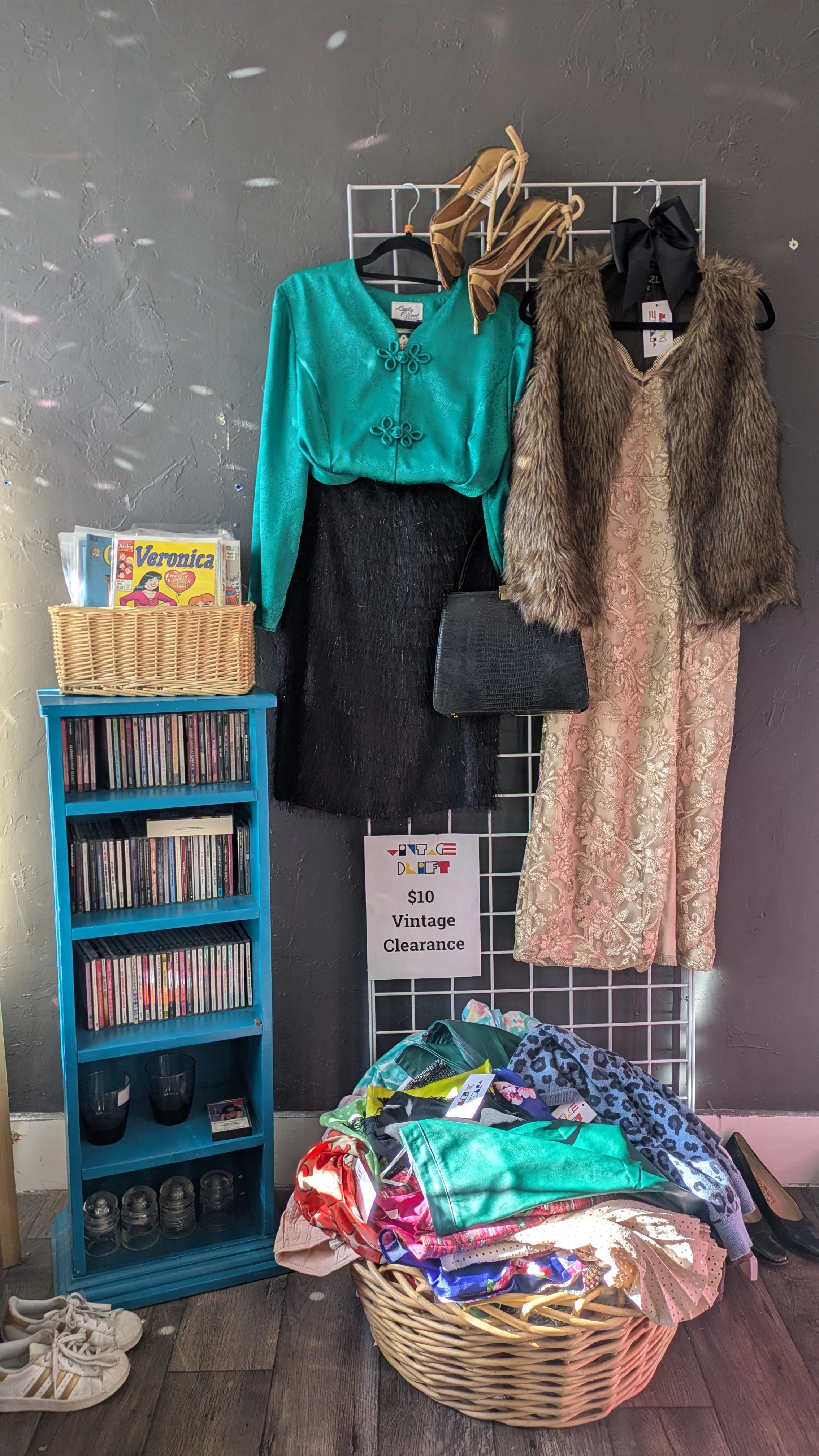 Vintage clearance display with hanging clothing and a basket of fabric, CDs, and shoes nearby.