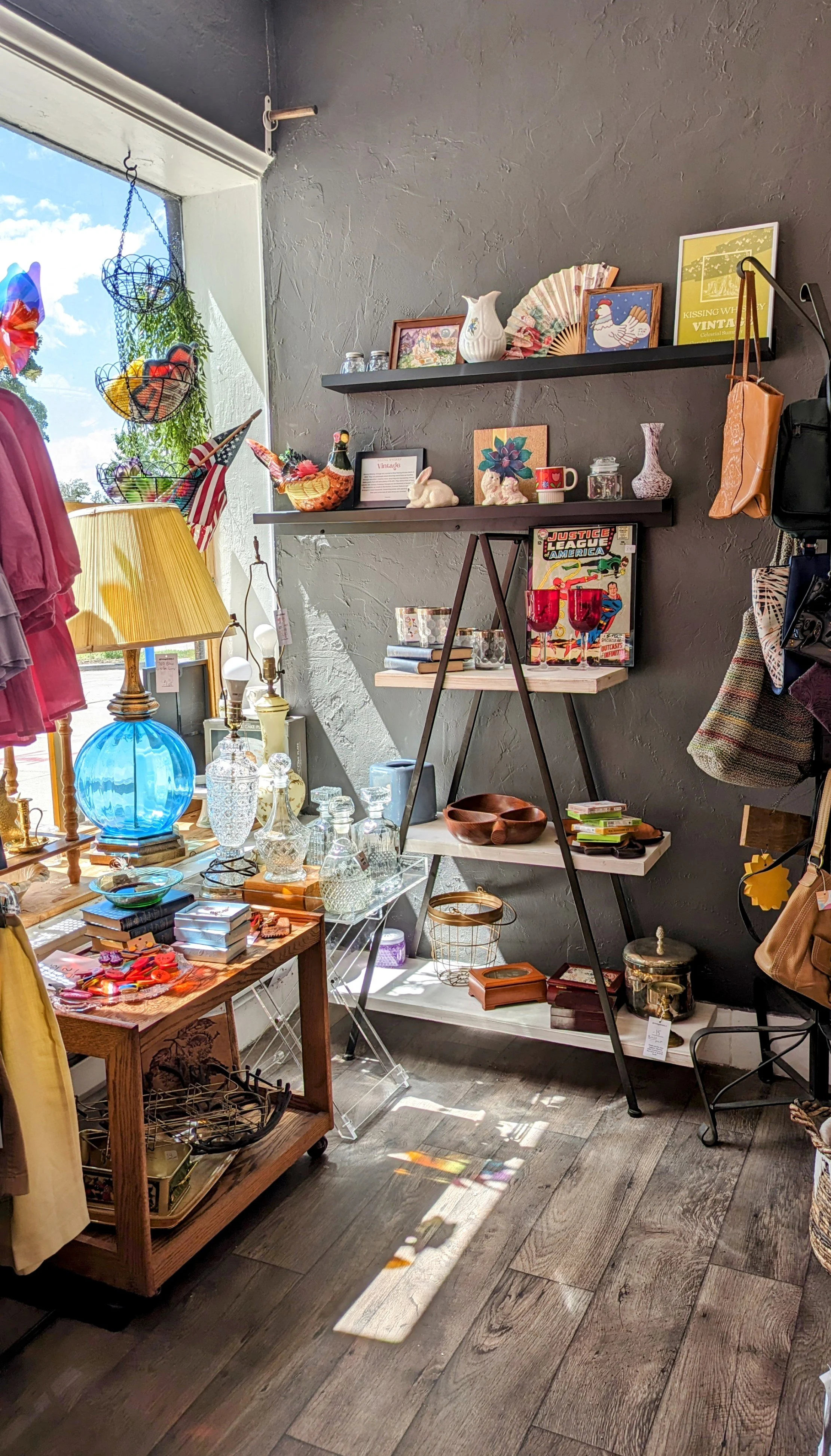 Interior of a vintage store with shelves and tables displaying glassware, books, jewelry, and decorative items. A window with sunlight and shadows cast on the wooden floor.