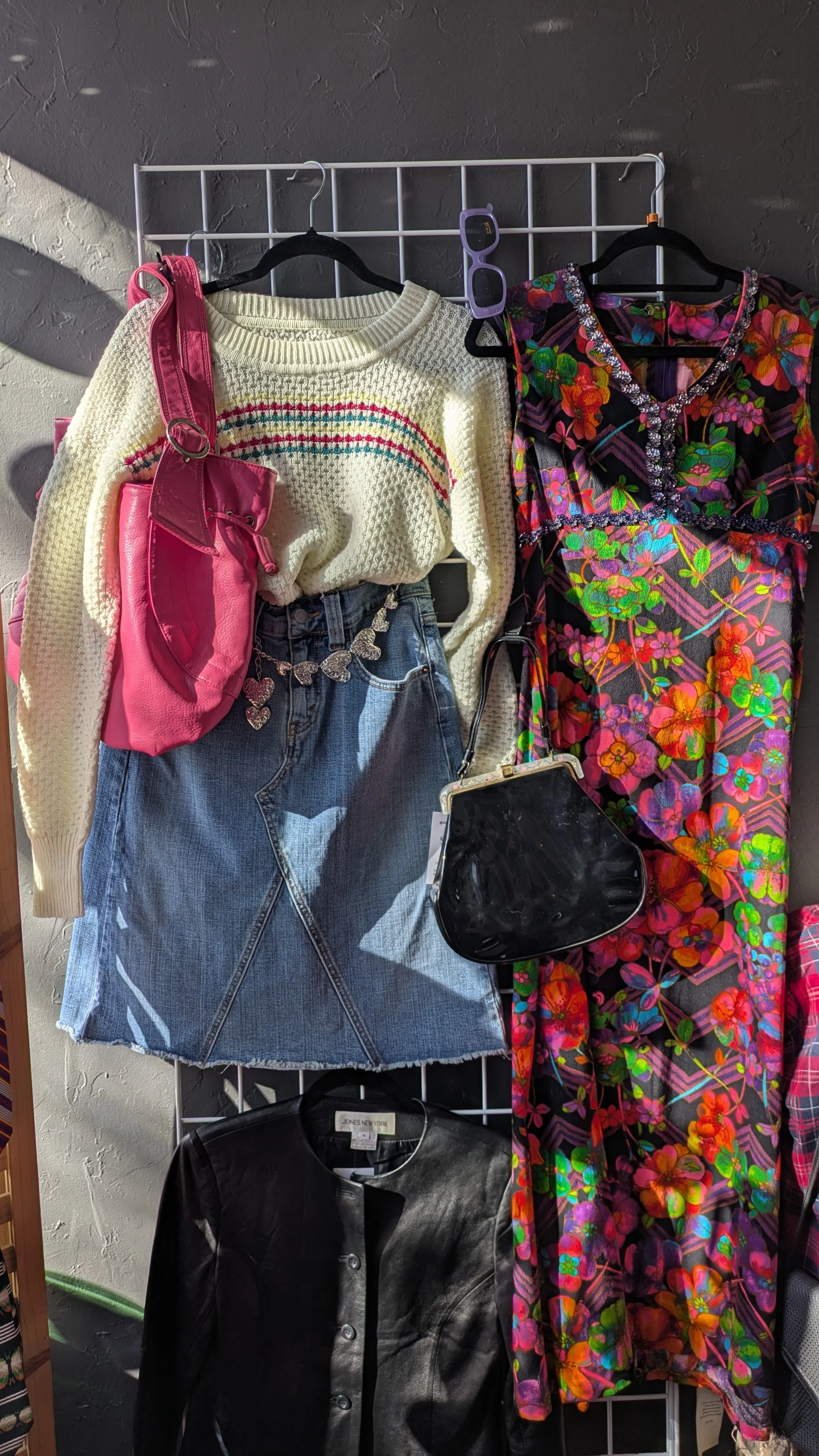 Display of colorful clothing and accessories, including a cream sweater with rainbow stripes, a denim skirt, a black leather jacket, a pink bag, a black purse, purple sunglasses, and a colorful floral dress.