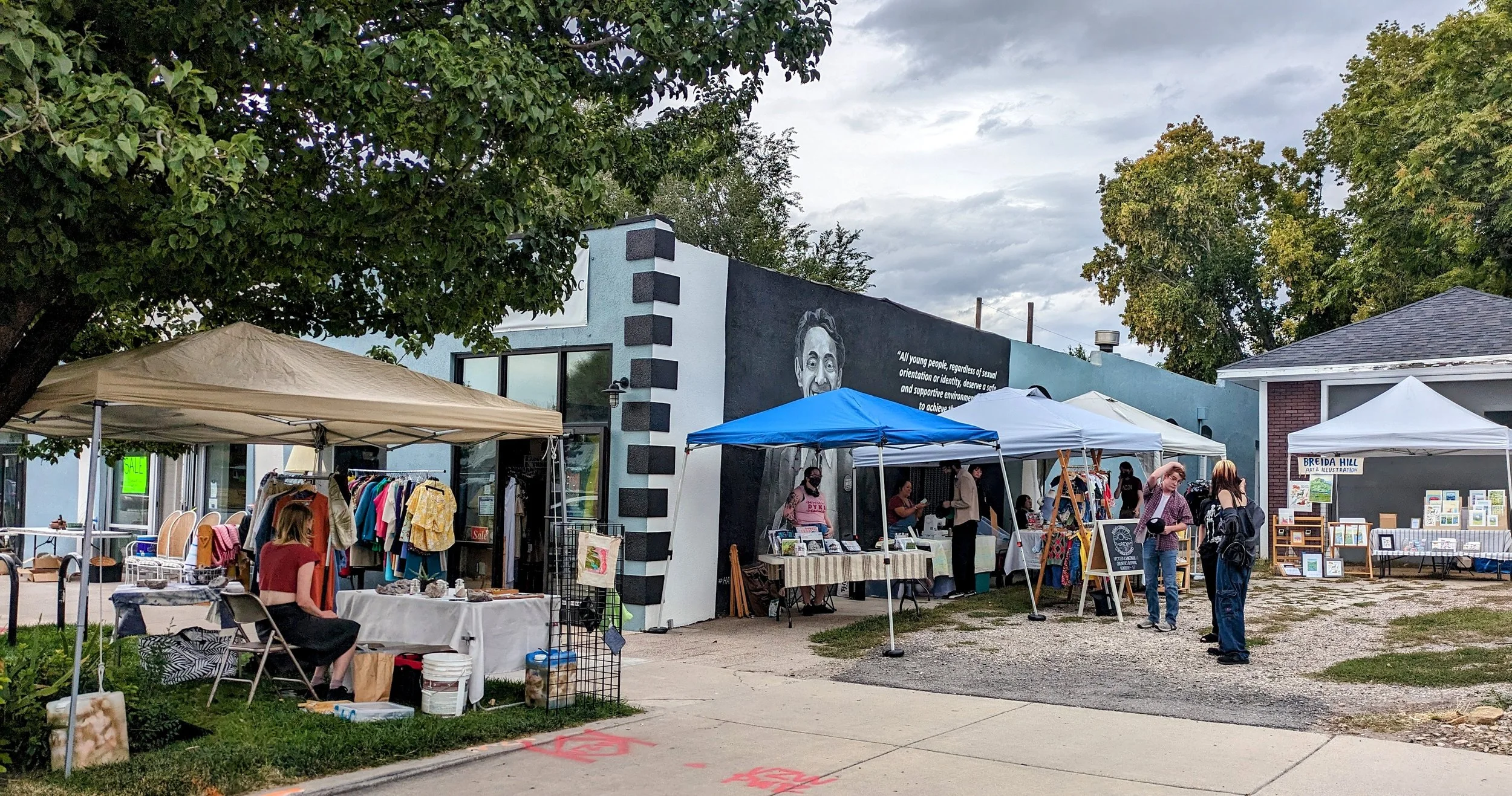 Outdoor art fair with vendor tents and people browsing on a sidewalk in front of a building with murals and trees.