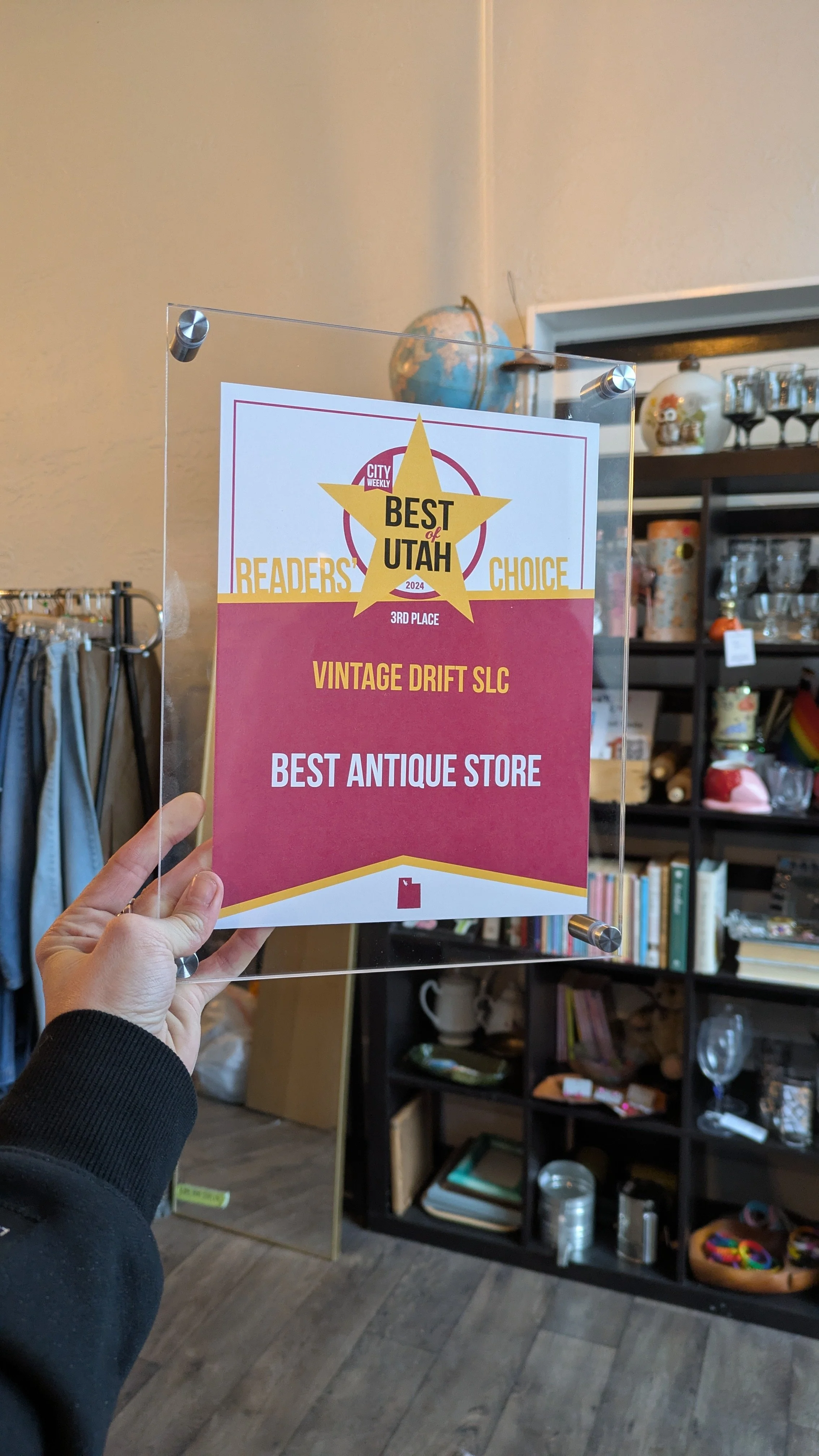 A person holding a clear acrylic award plaque that recognizes the store 'Vintage Drift SLC' as the third place winner in the 'Readers' Choice' for the 'Best Antique Store' in Utah. The background shows a store interior with shelves of various items.