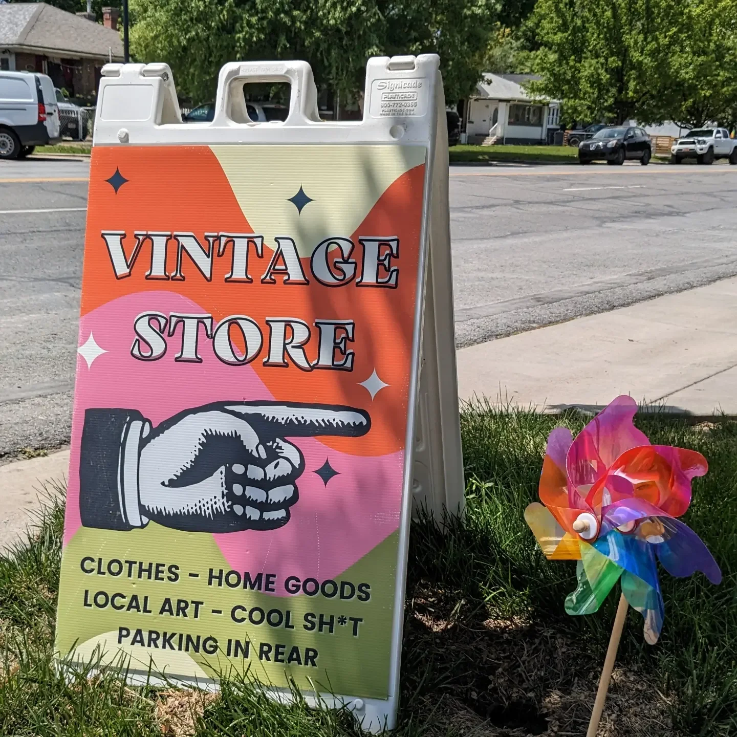 A/crosswalk sign advertising a vintage store with a pointing hand illustration, on a grassy area next to the street. It lists items like clothes, home goods, local art, and parking behind the store. A colorful pinwheel is nearby.