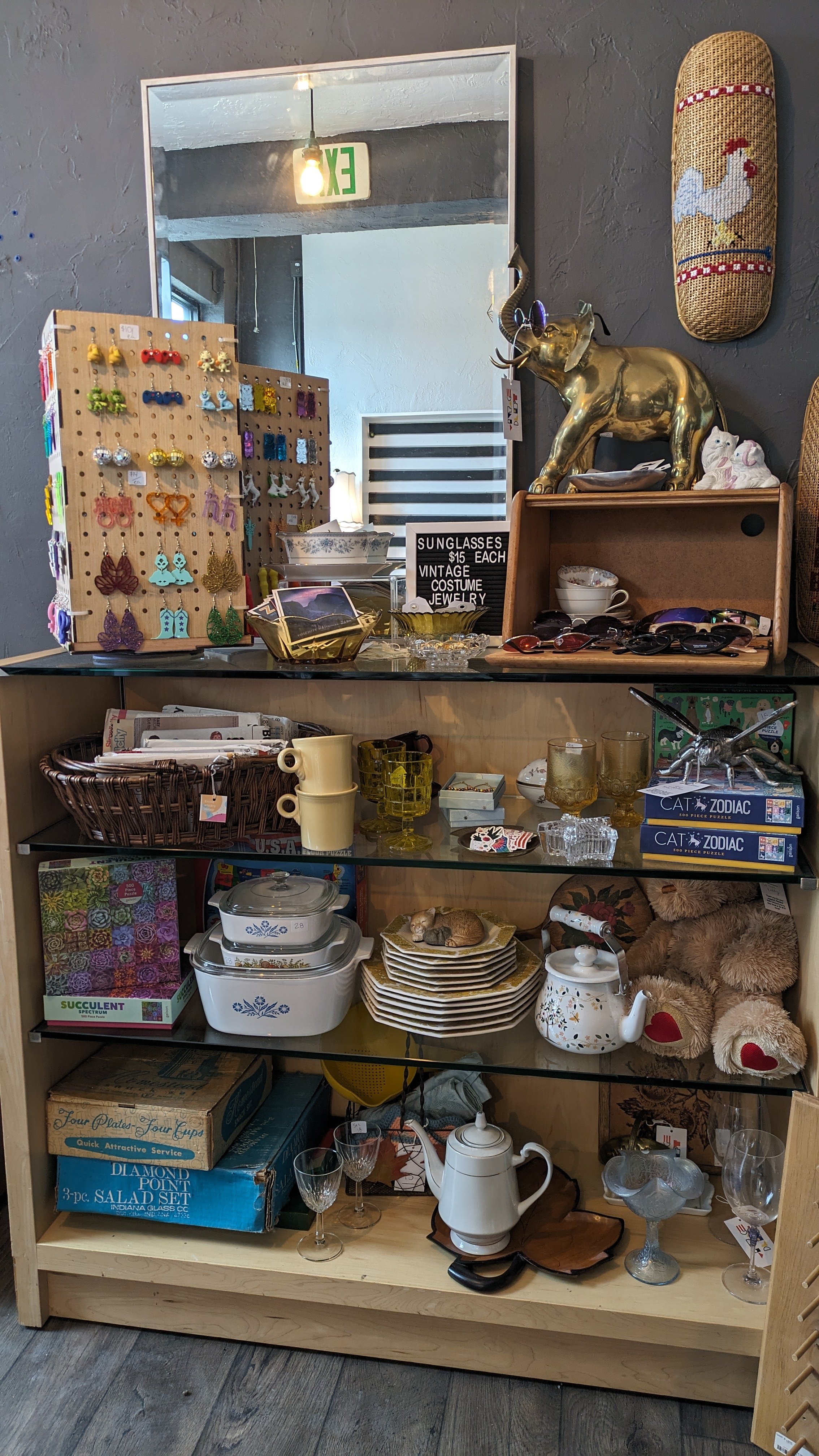 A wooden shelf displaying various vintage and decorative items, including glassware, dishware, toys, jewelry, and small collectibles, with a mirror and wall decorations in the background.