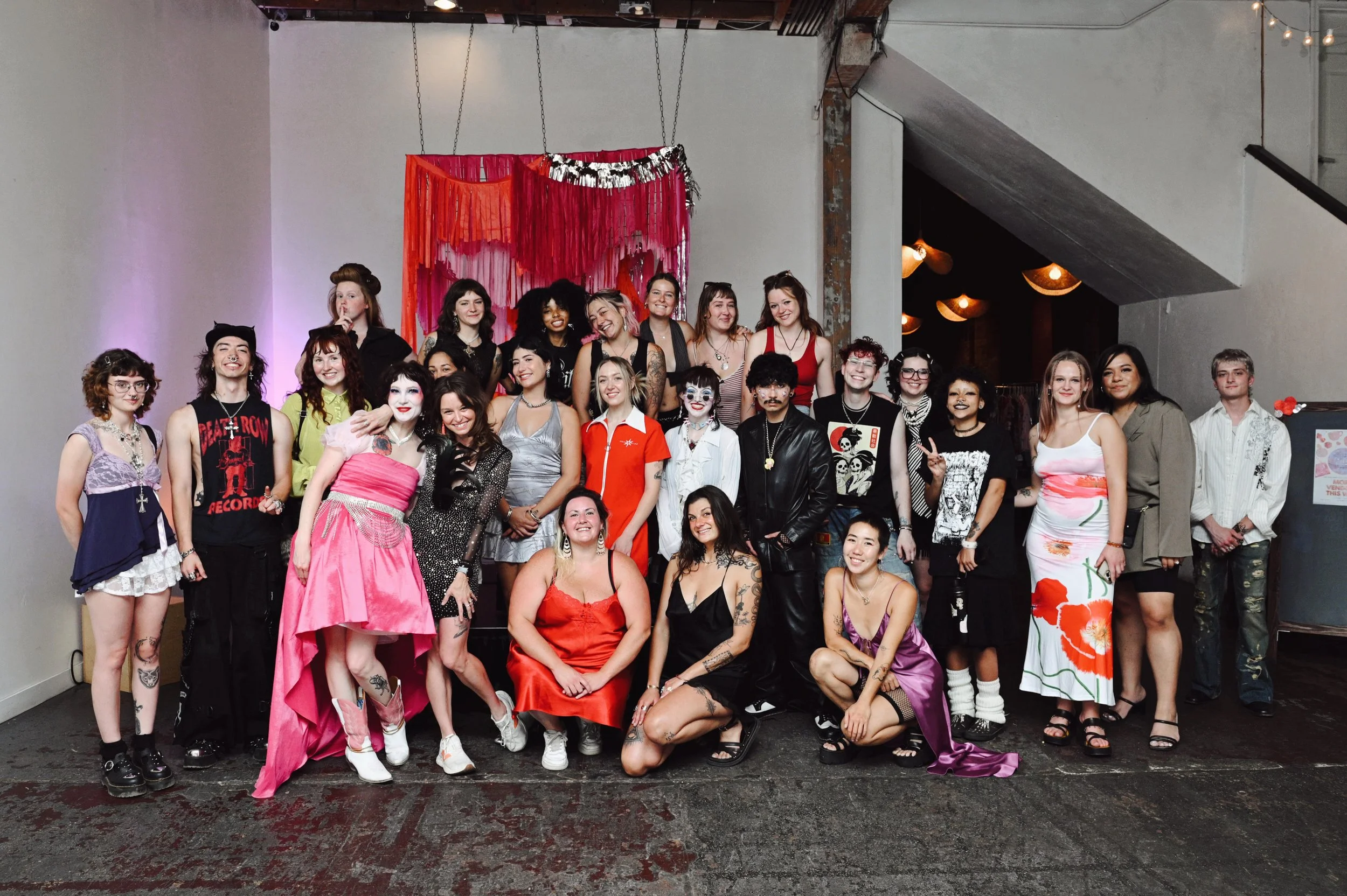 Group of diverse people dressed in various costumes and outfits, posing together indoors in front of a colorful backdrop with red and pink fabric decorations.