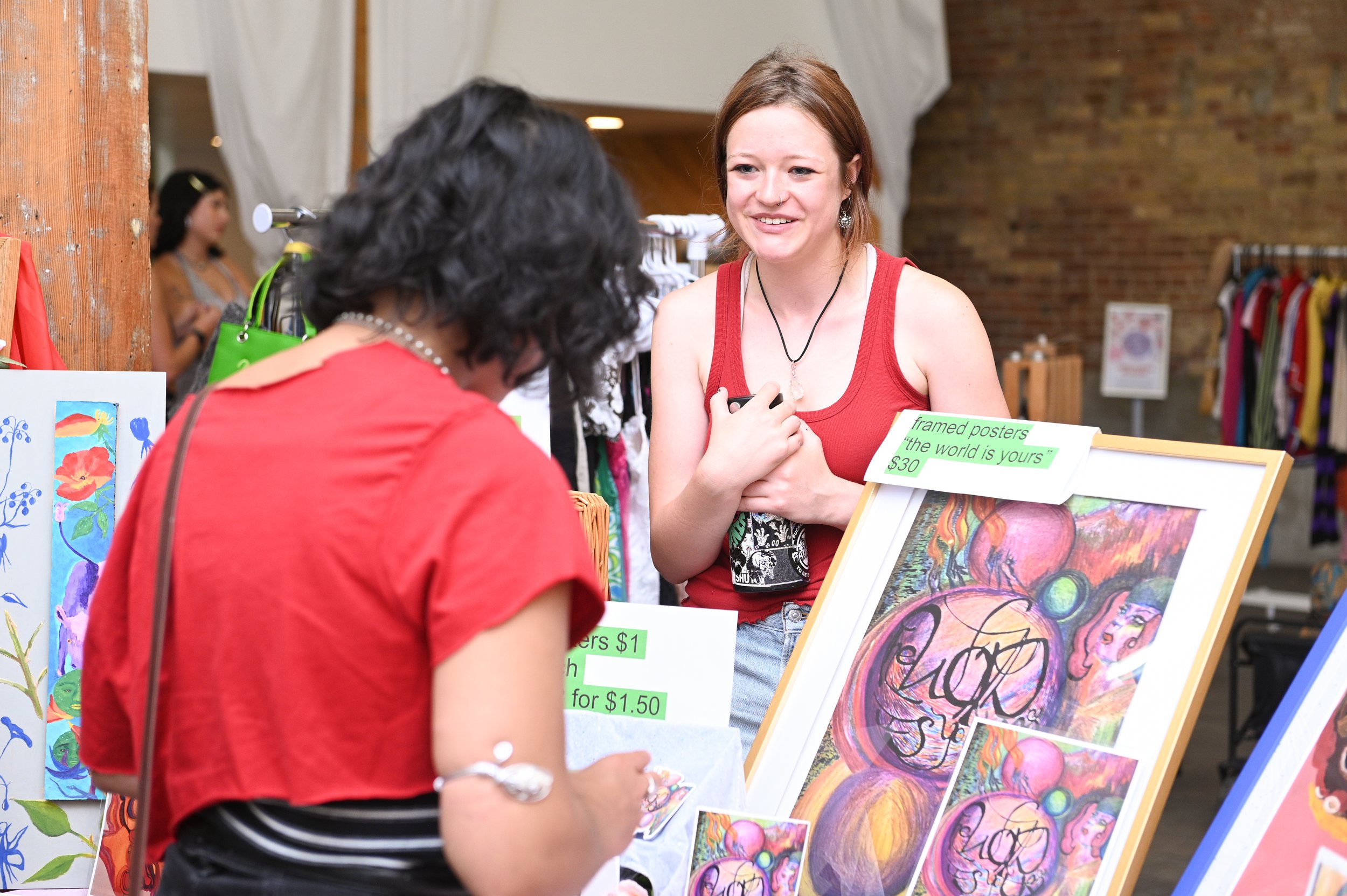 A woman with red hair in a red tank top sells framed art and posters at a market stall, talking to a customer who is wearing a red top and striped skirt. The stall displays colorful artwork including abstract and floral designs, with price signs for the art and posters.