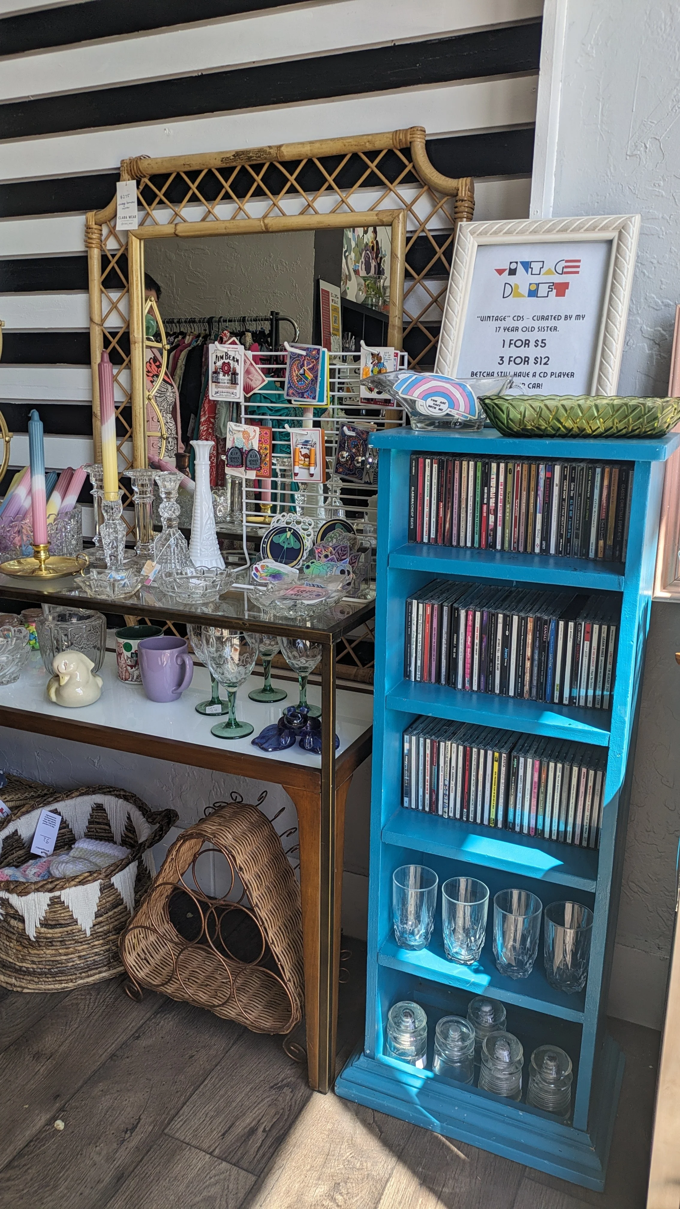A vintage thrift store display with a blue wooden CD shelf, glassware, candles, decorative items, jewelry, and clothing.
