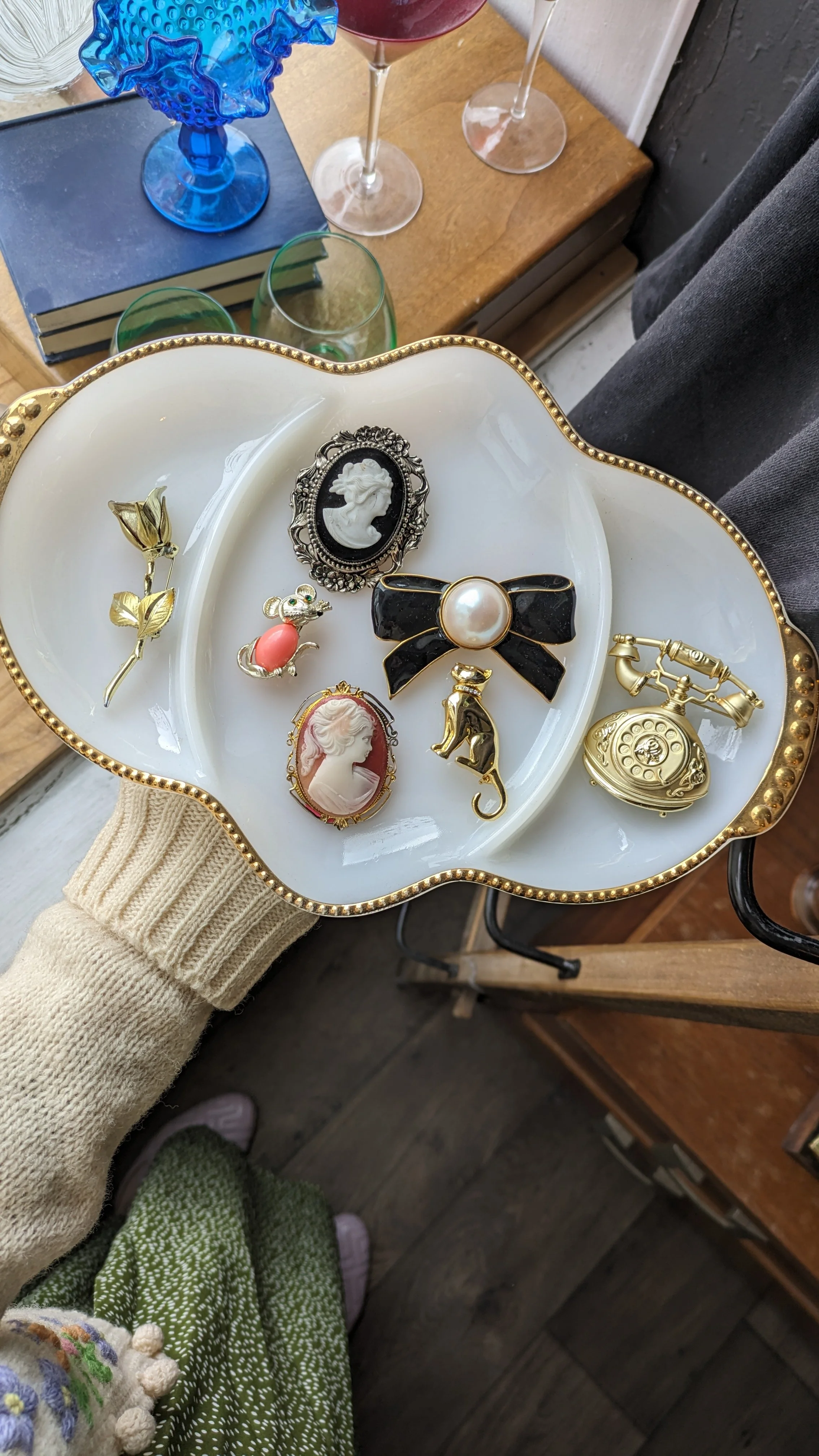 A white scalloped tray with gold trim holding vintage-style jewelry including brooches, pins, and a tiger-shaped pin, held by a person wearing a beige sweater.