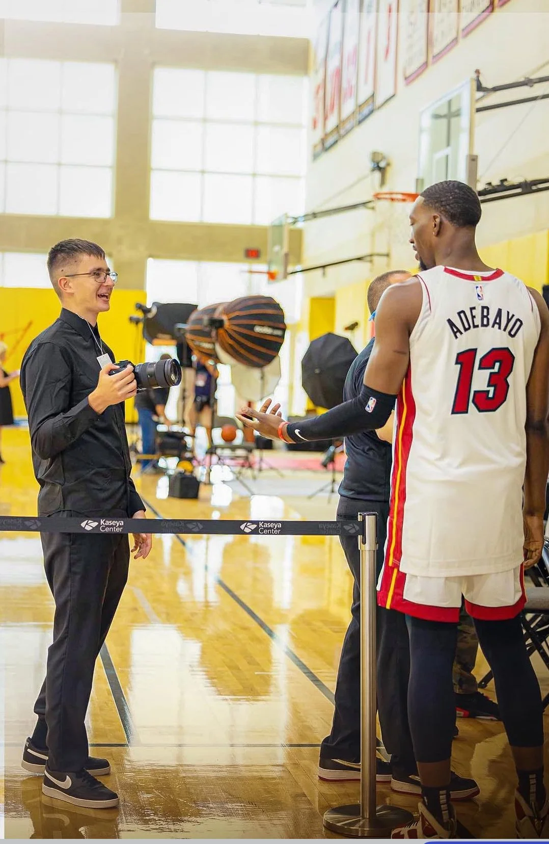 A photographer talks to a basketball player wearing a white and red jersey with the name 'Adebayo' and the number 13, inside a gymnasium during a media event. The basketball player is tall with dark skin and short hair, and stands near a barrier with the label 'Kaseya Center.' In the background, there are photography lights, basketballs, and other people.