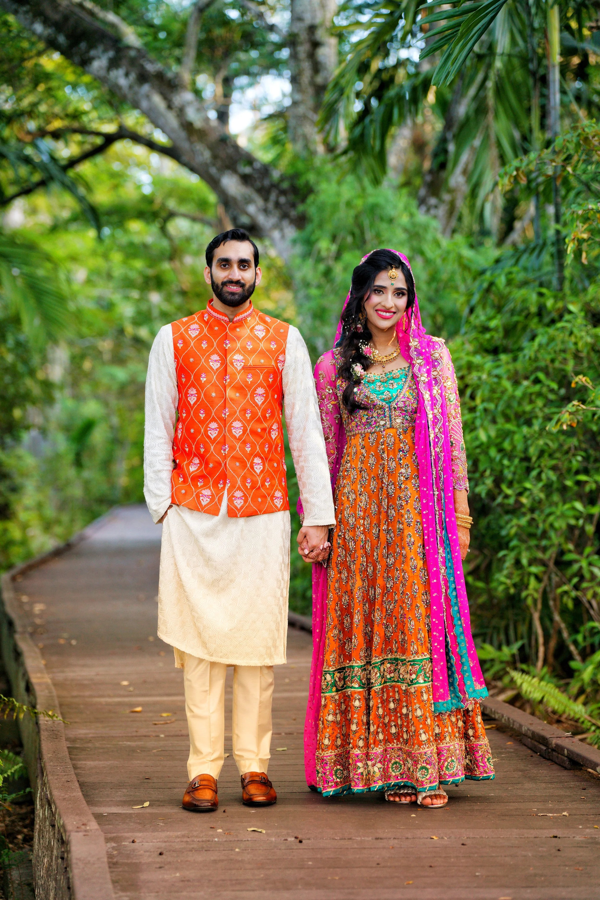 A couple dressed in traditional Indian attire standing hand-in-hand on a wooden pathway surrounded by lush green trees.